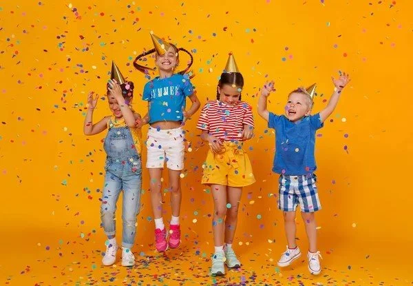 Four children in party hats celebrate, jumping and smiling as colorful confetti falls, against a bright yellow background.
