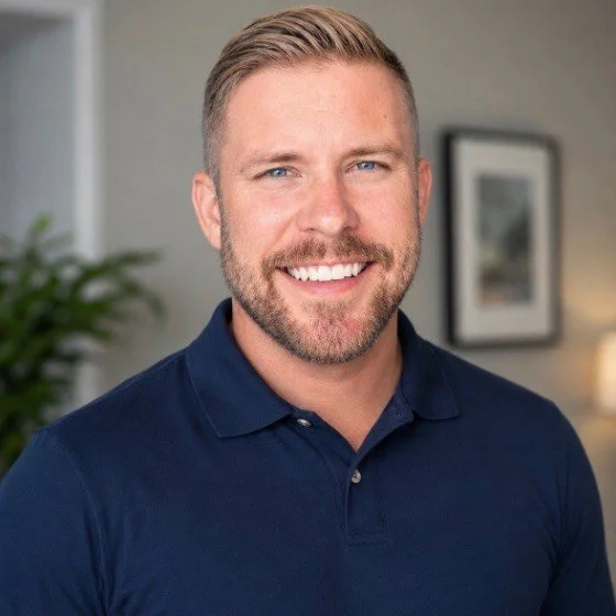 Smiling man therapist with short blond hair and beard, wearing a navy polo shirt, stands indoors with blurred background.