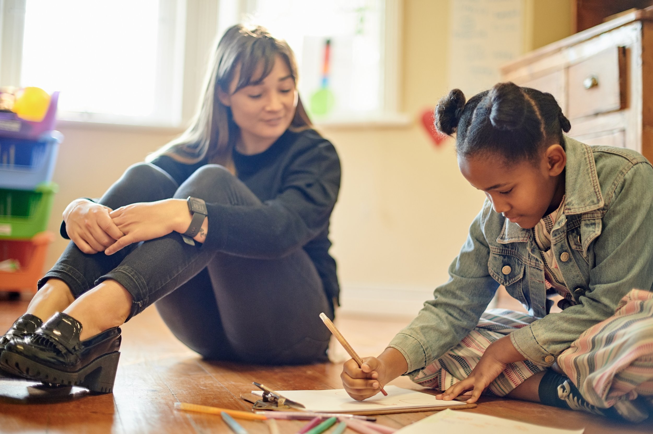 A woman and a girl are sitting on the floor, engaging in drawing with colored markers, surrounded by art supplies in a cozy room.