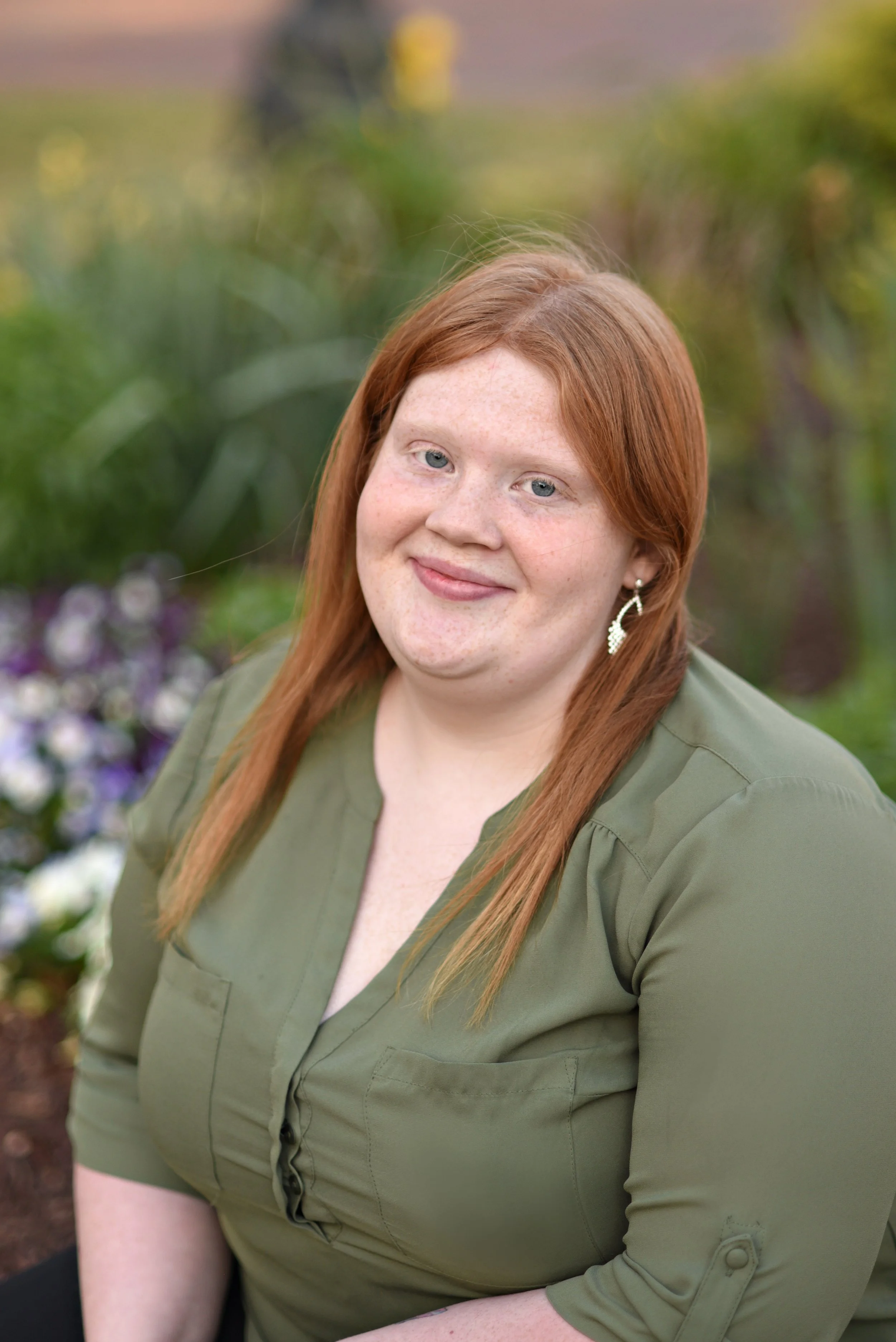 Woman therapist with long red hair and green blouse smiles outdoors with flowers and greenery in the background.