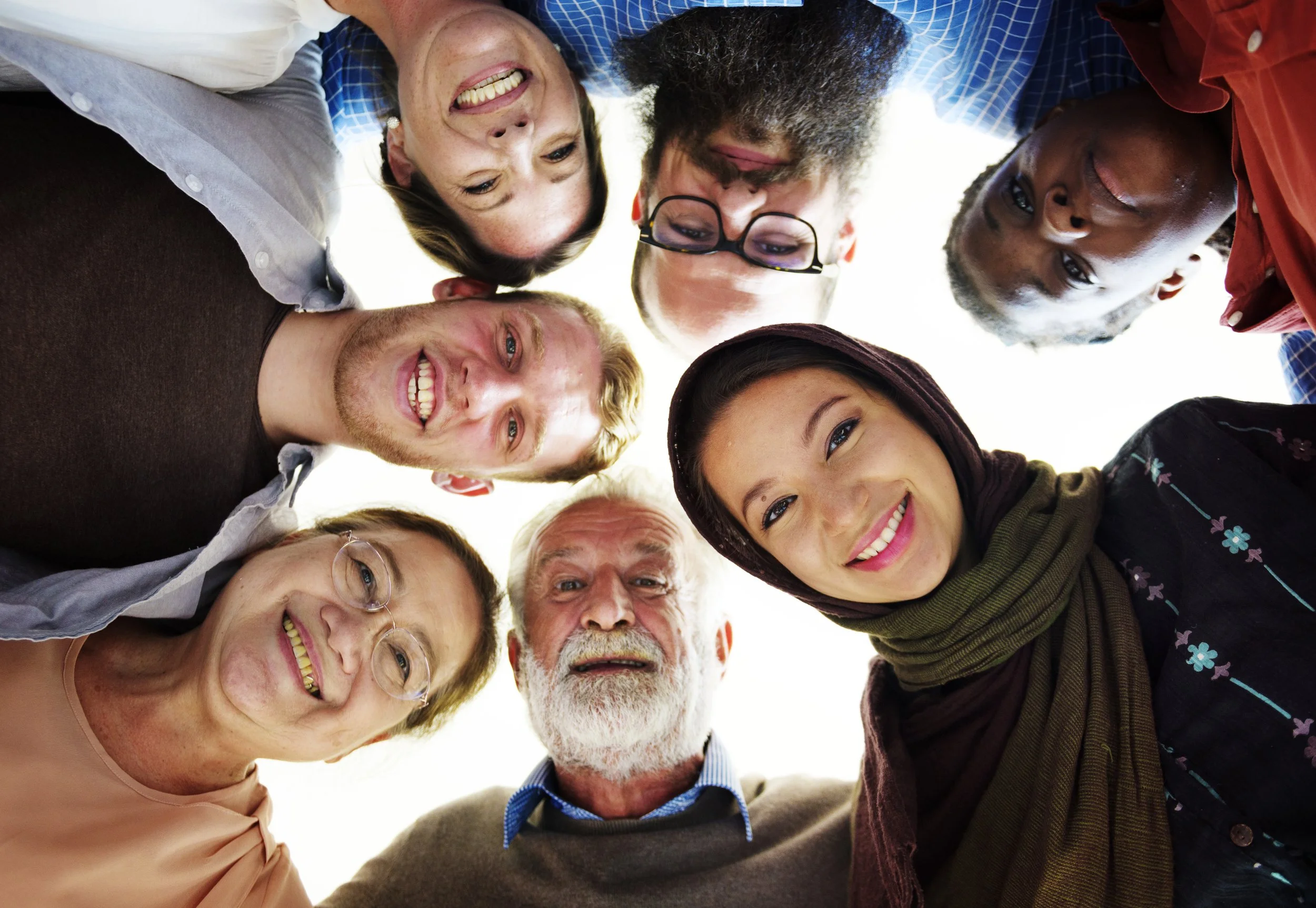 Seven diverse people stand in a circle, smiling and looking down at the camera from above, forming a close group.