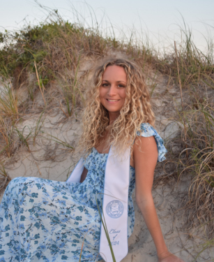 Therapist in a blue floral dress and graduation stole sits on sandy dunes with tall grass, smiling at the camera.
