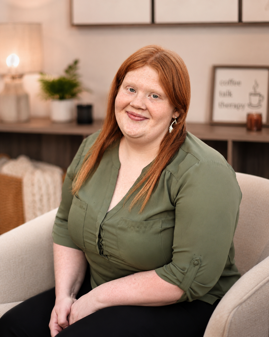 Female therapist with red hair and a green blouse sits smiling in a cozy, well-lit room with warm decor.