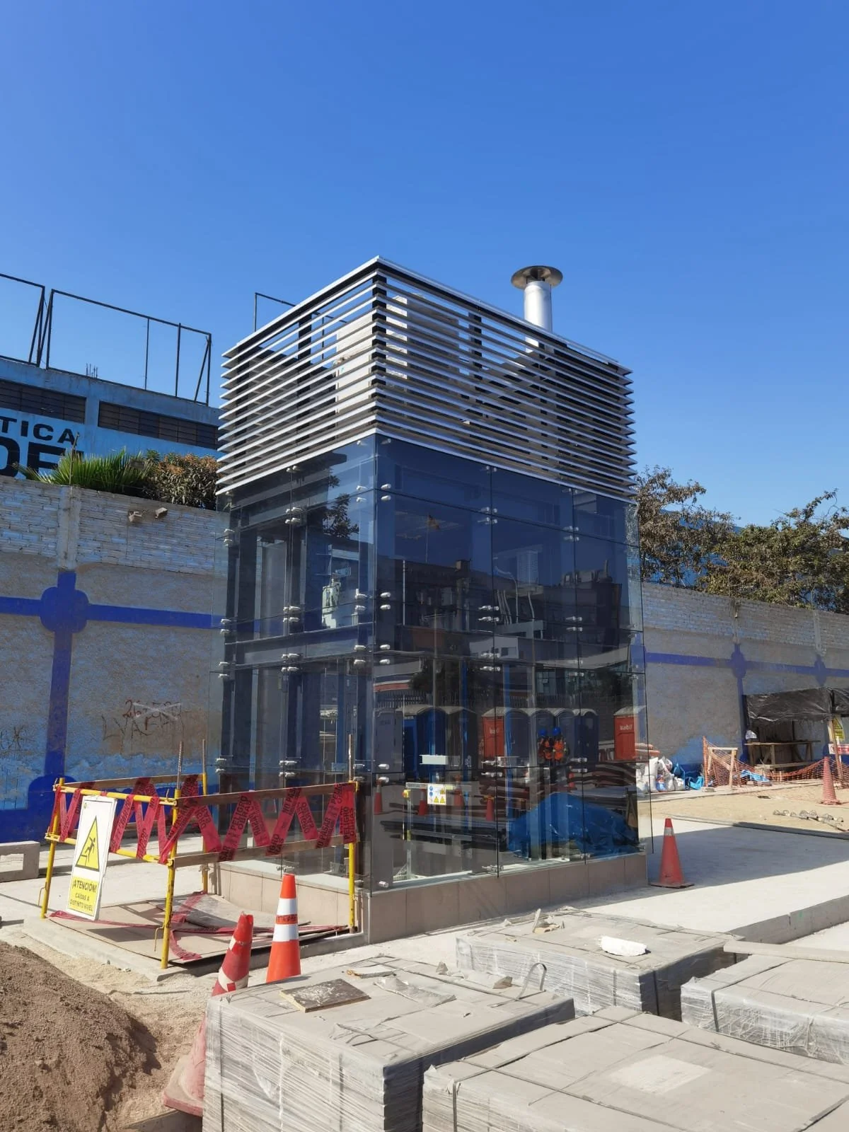 Estructura de elevador exterior de cristal en construcción, rodeada de conos de seguridad y vallas, en un día soleado.