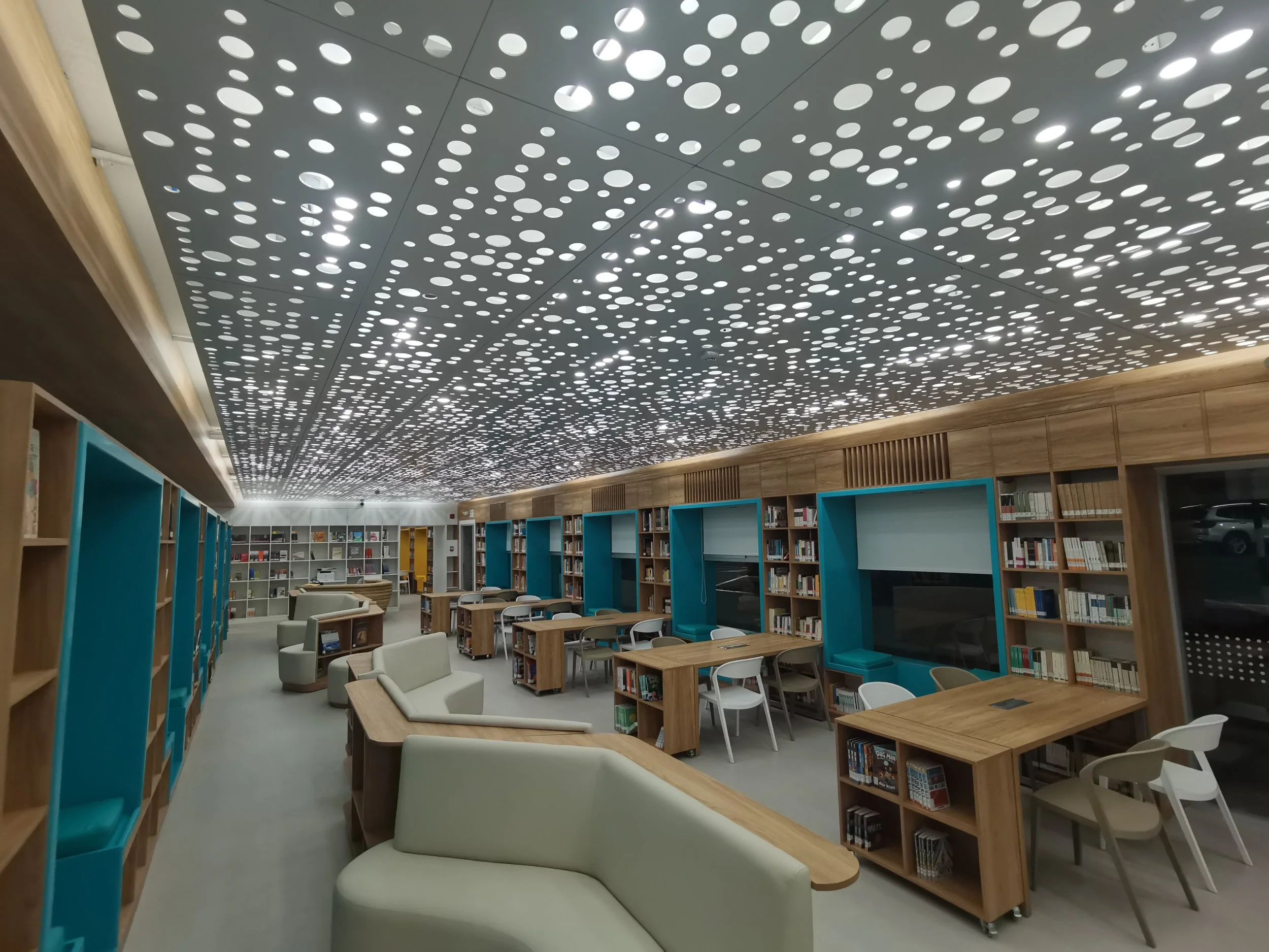 Biblioteca moderna con estanterías de madera, mesas de estudio y sillas blancas, techo con diseño de círculos blancos y luz brillante.