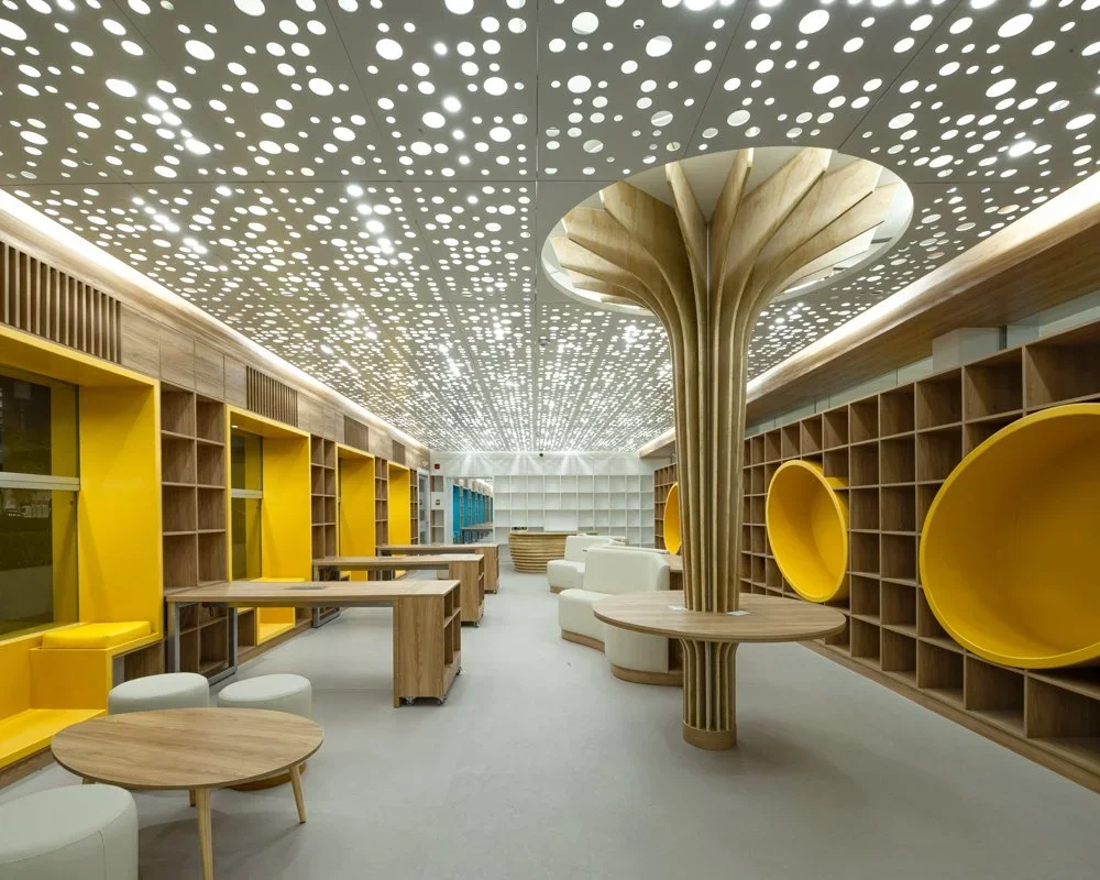 Interior moderno con estanterías de madera, ventanas con marcos amarillos, sillones blancos y taburetes, techo decorativo con círculos blancos y estructura central de madera en forma de árbol.