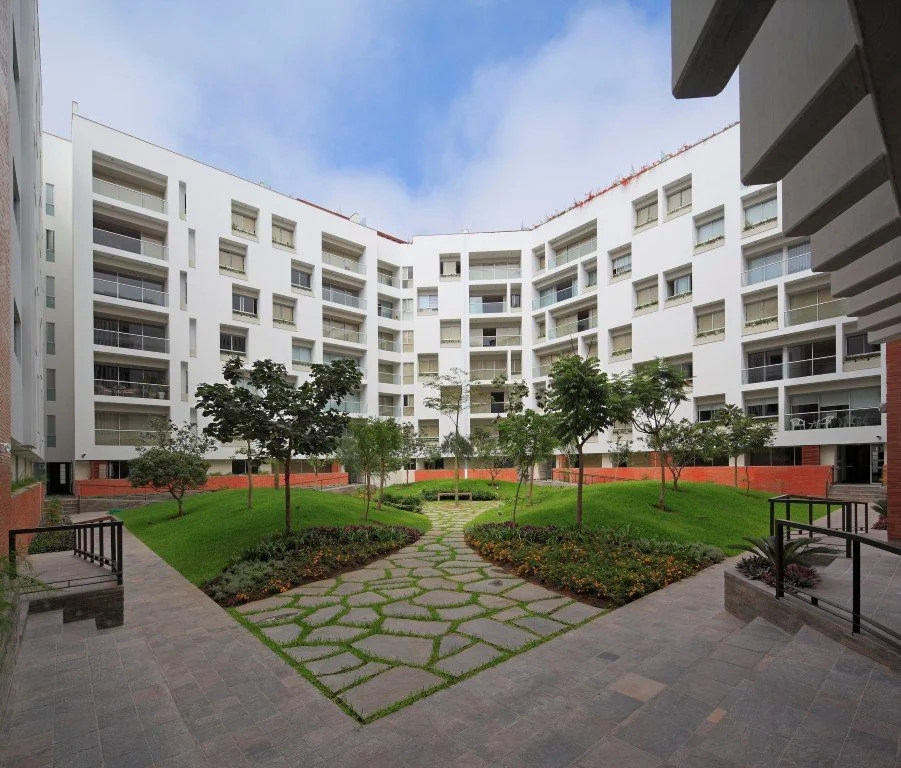 Vista de un jardín en el centro de un conjunto habitacional de varios pisos con áreas verdes, árboles y un camino de piedra.