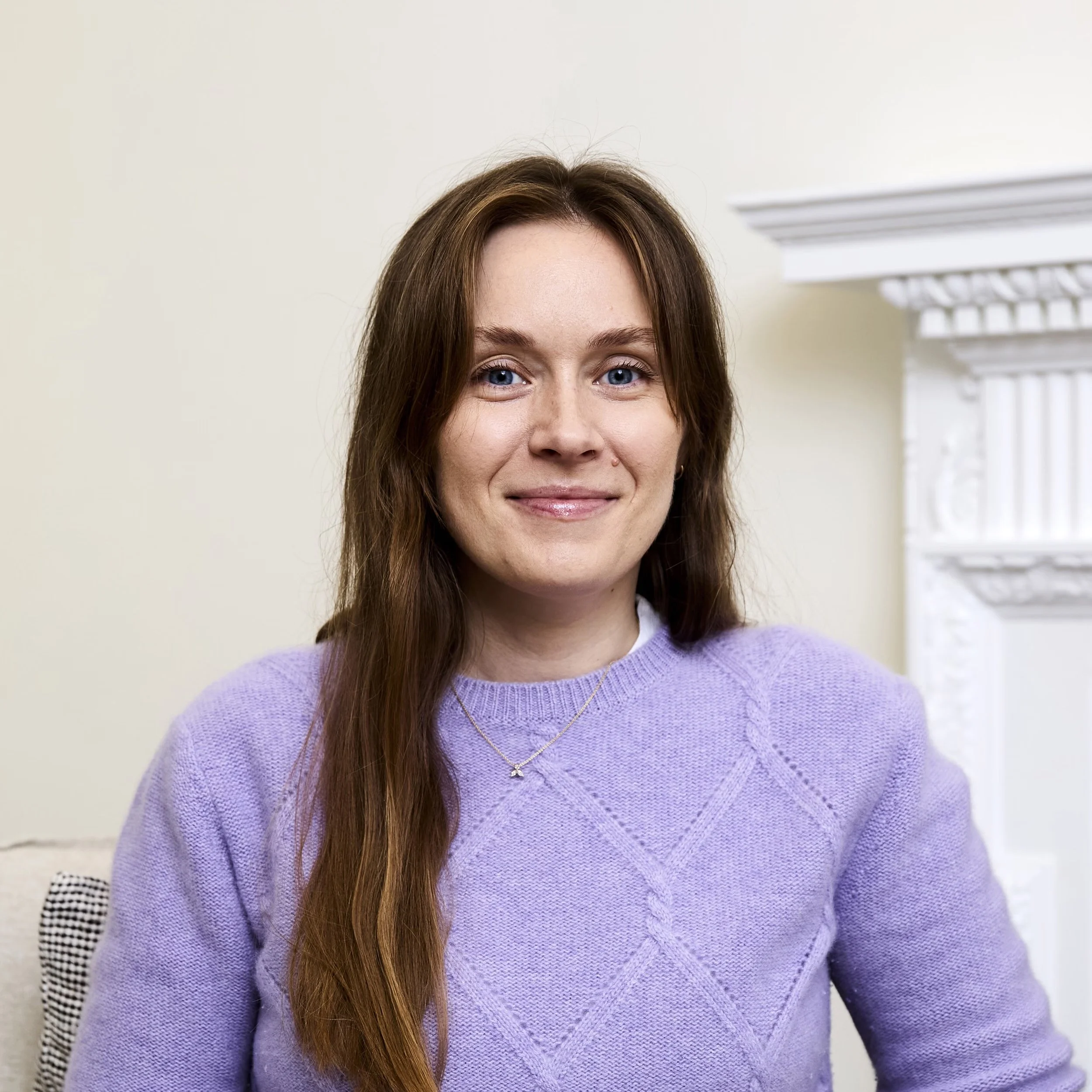 Portrait of a young woman with long brown hair, blue eyes, smiling, wearing a lavender sweater, and a gold necklace, sitting indoors with light-colored walls and a white ornate fireplace in the background.