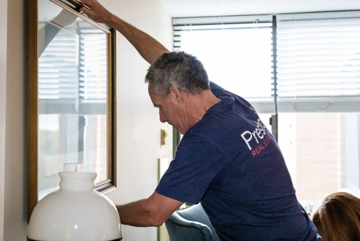 A man cleaning a framed mirror in a room with large windows and blinds.