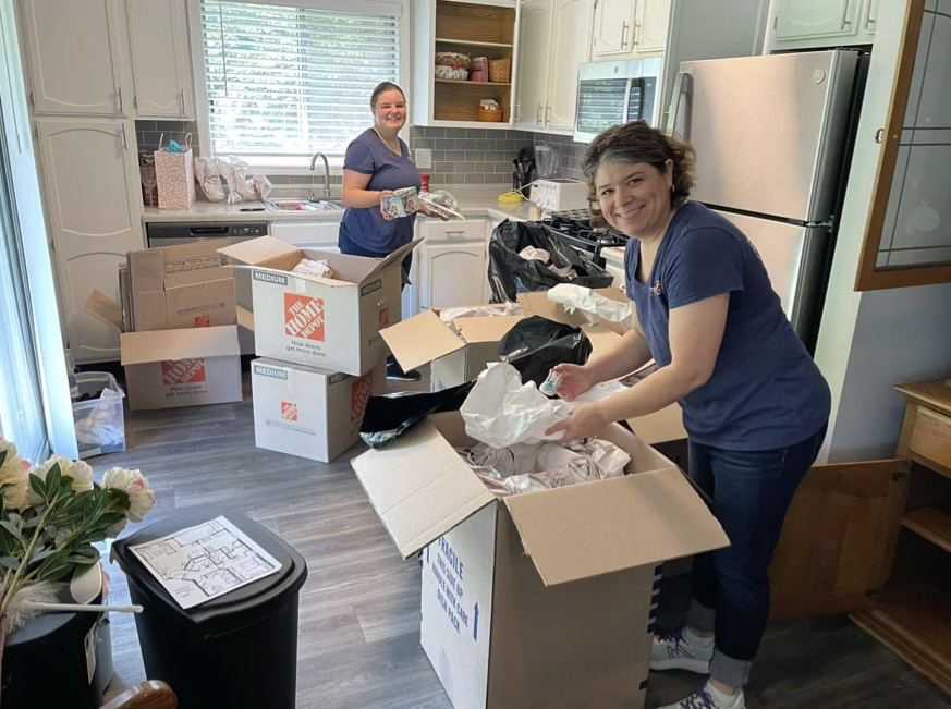 Two women packing boxes in a kitchen with sunlight coming through a window.