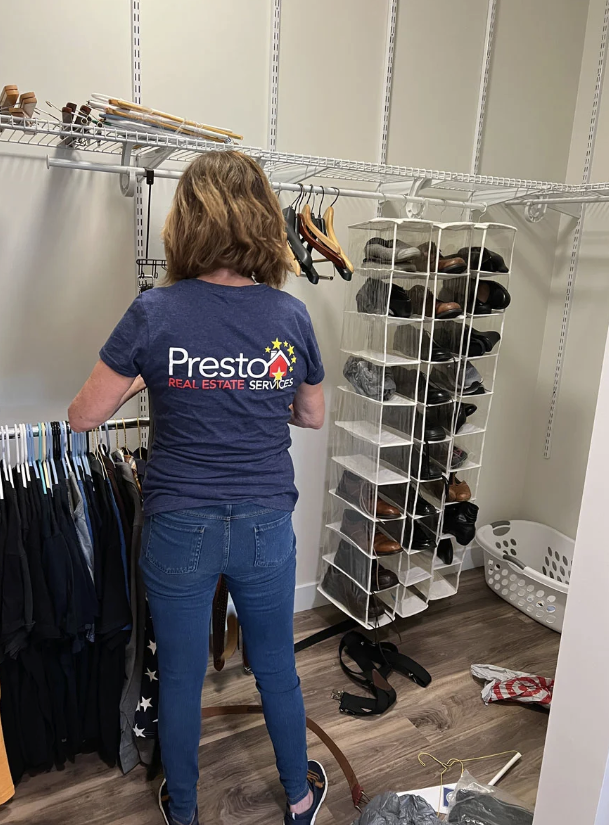A woman with shoulder-length hair wearing a navy blue Presto real estate services t-shirt and blue jeans organizing clothes in a closet with hanging rods, a shoe rack, a laundry basket, and various accessories.