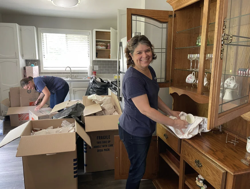 A woman smiling while packing dishes into a cabinet in a kitchen, with a man in the background organizing boxes.