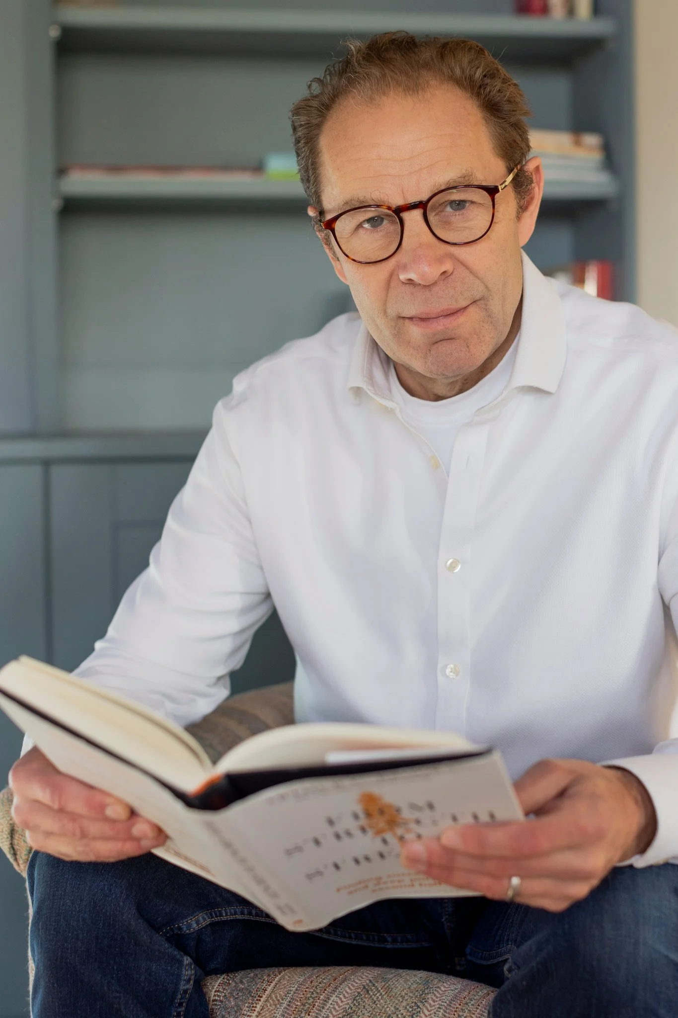 A man wearing glasses and a white shirt sitting on a chair and reading a book.