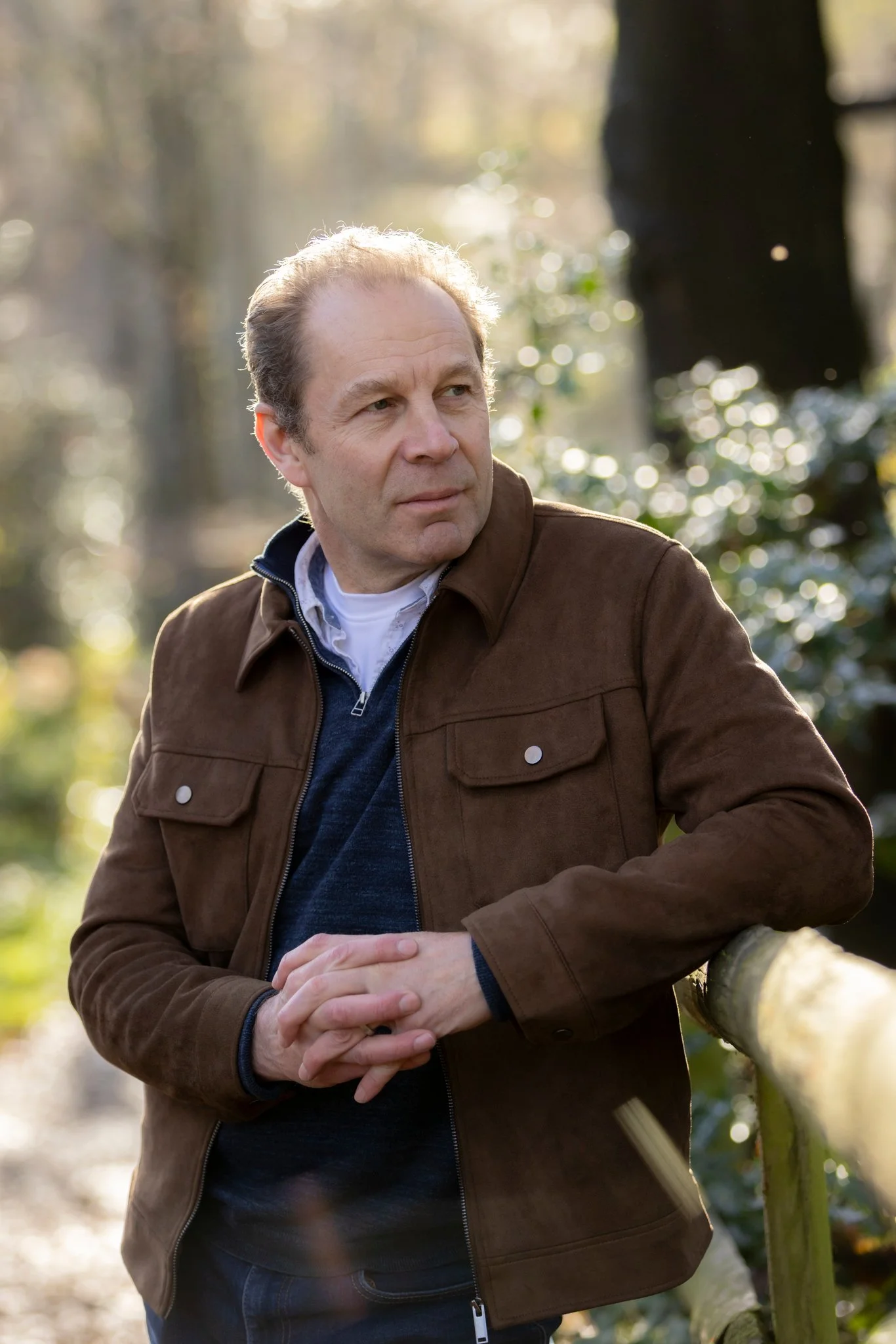 A middle-aged man with light skin and short, wavy hair standing outdoors on a sunny day in a wooded area. He is wearing a brown jacket over a white shirt and a blue sweater. He is leaning on a wooden railing with his hands clasped, looking thoughtfully to the side.