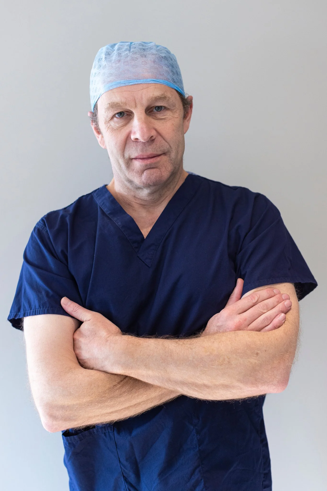 A middle-aged male healthcare professional wearing navy blue scrubs and a light blue surgical cap, standing with arms crossed and looking directly at the camera against a plain light background.
