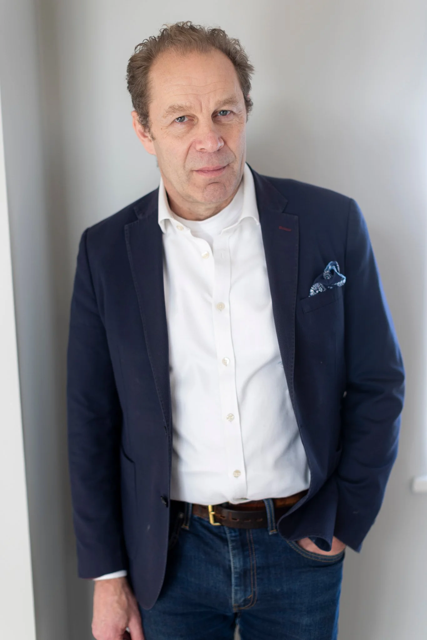 A middle-aged man with light skin, brown hair, and blue eyes is standing indoors against a plain light-colored wall. He is wearing a dark blazer, white dress shirt, and jeans, with one hand in his pocket and a serious expression on his face.