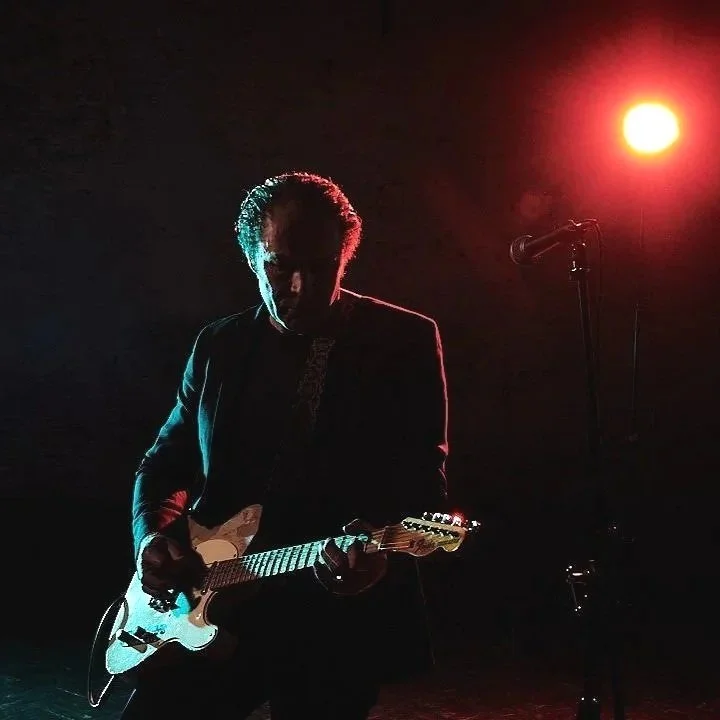 Charles Knowles plays electric guitar on stage, silhouetted against red and blue lighting with a microphone nearby.