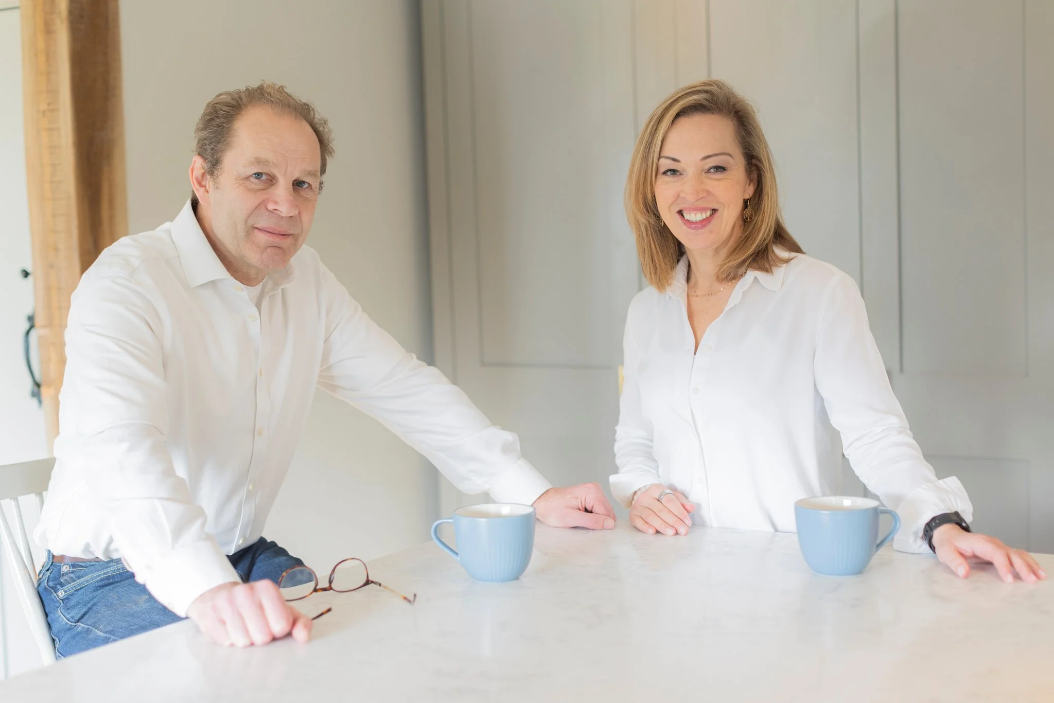 A man and a woman standing at a kitchen counter with two coffee mugs, both wearing white shirts, smiling, with glasses resting on the counter.
