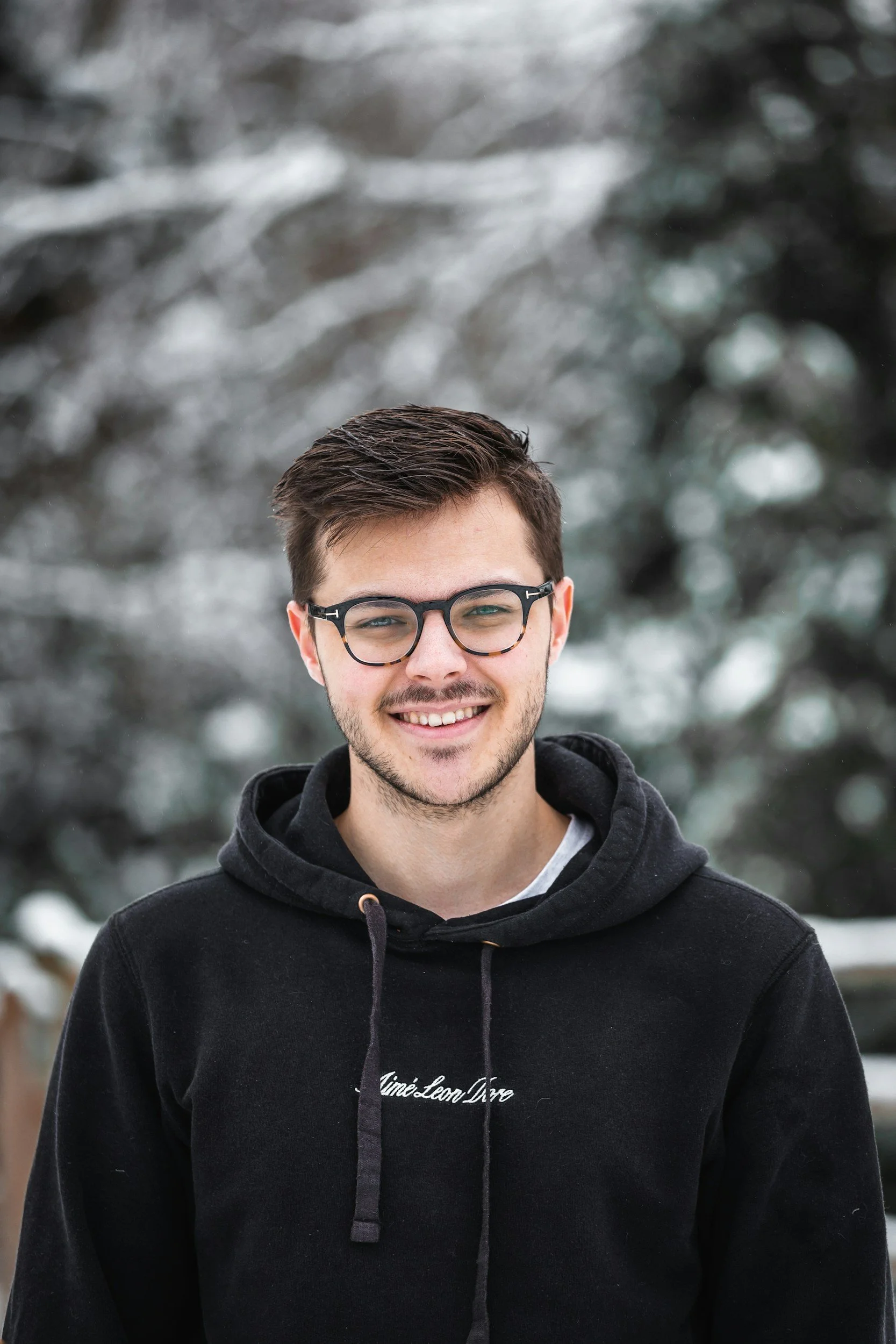 A young man wearing glasses and a black hoodie with white text, smiling outdoors with snow-covered trees in the background.