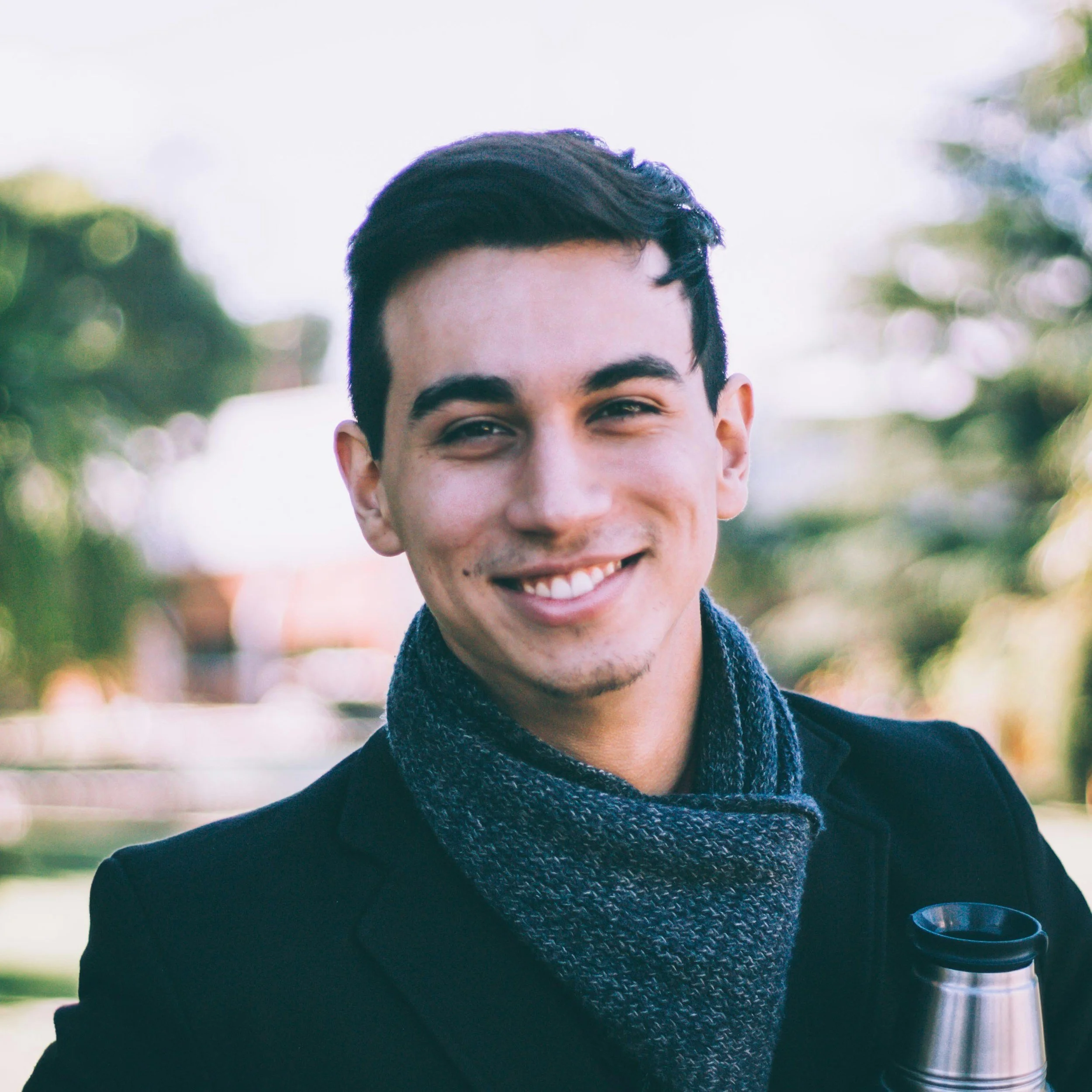Smiling young man with dark hair, wearing a dark coat and a gray scarf, holding a thermos outdoors with trees and blurred background.