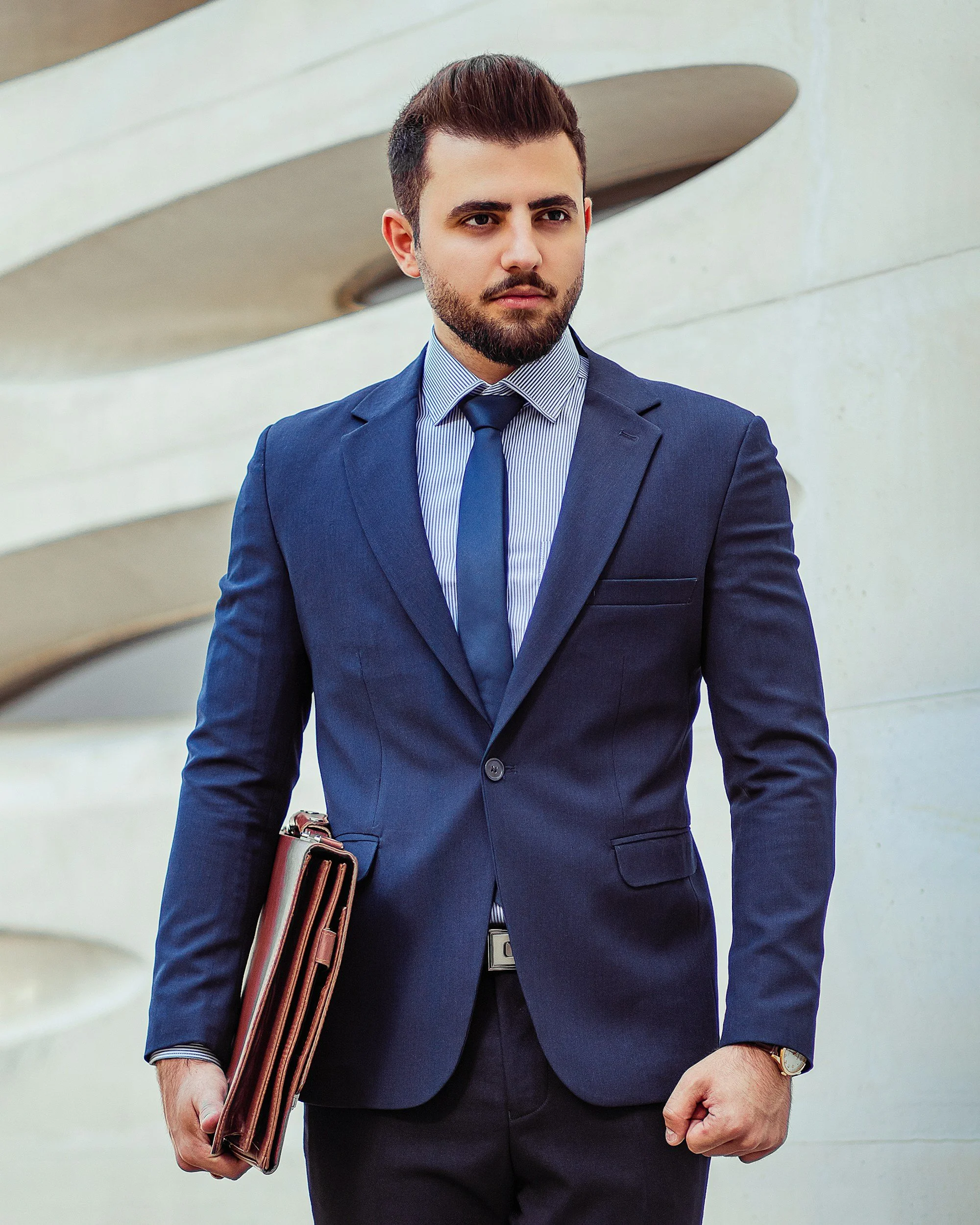 A man in a navy blue business suit holding a folder, standing outdoors, with modern architecture in the background.