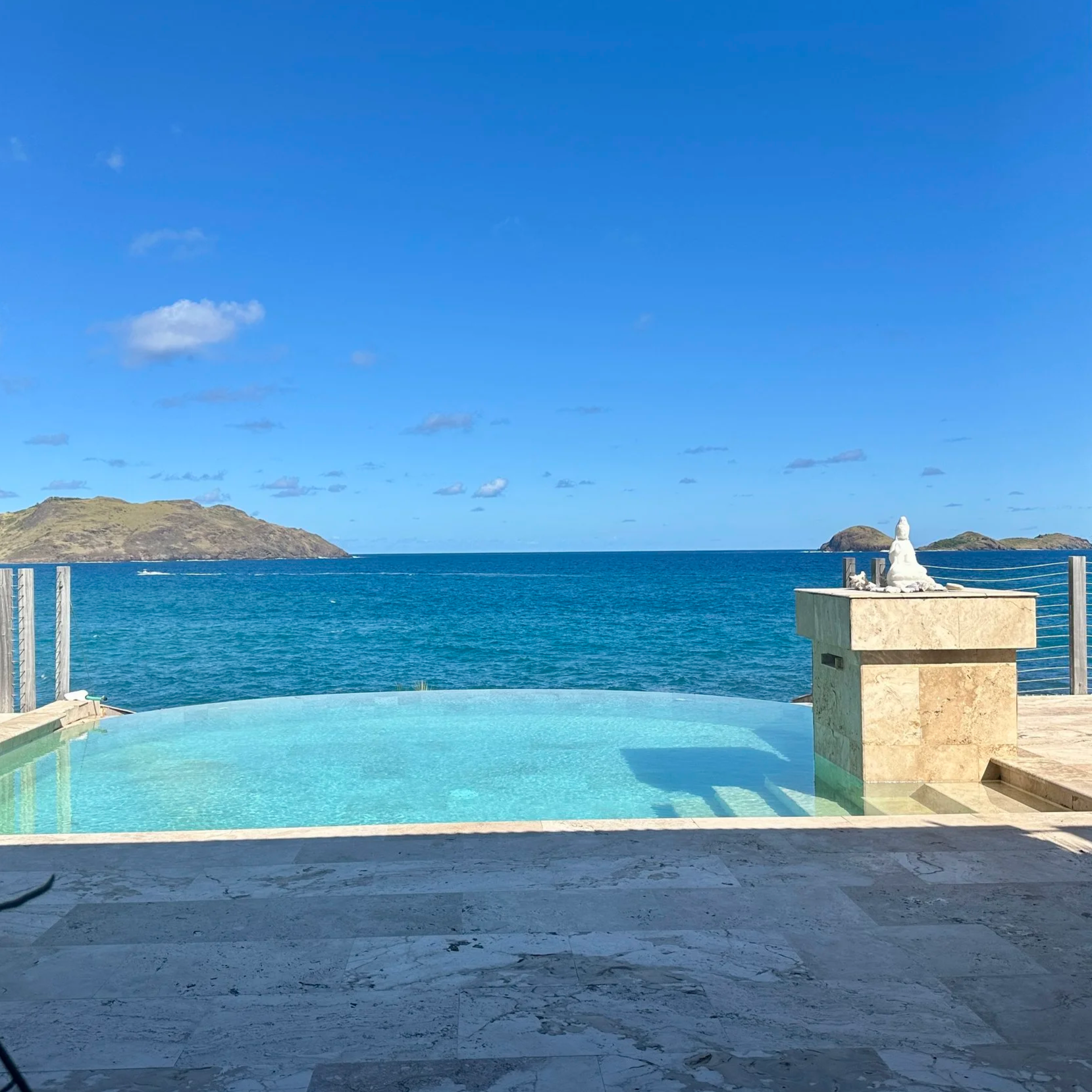 View of a swimming pool overlooking the ocean with islands in the distance, a clear blue sky, and a small white sculpture on a stone pedestal.