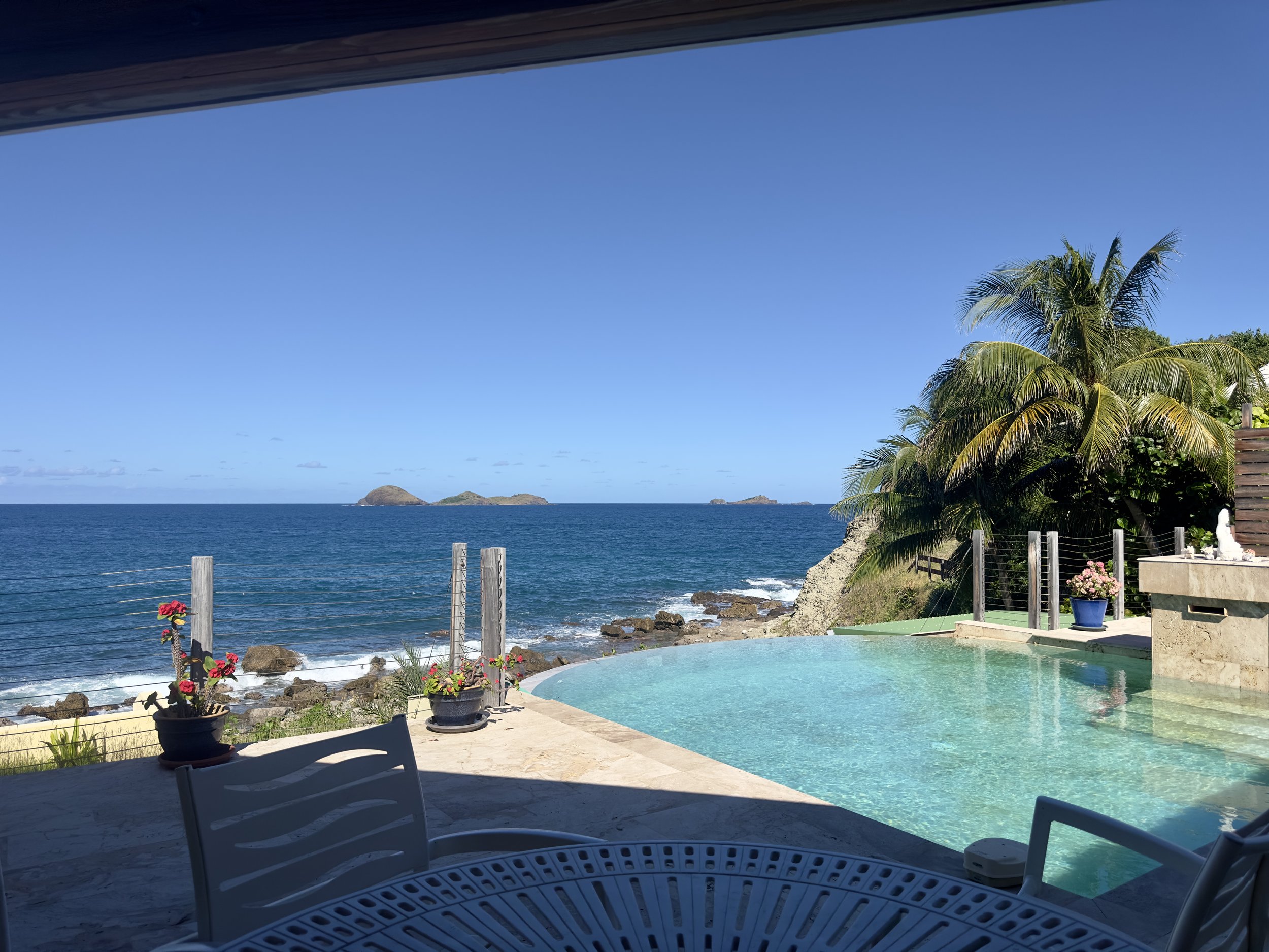 A view of the ocean with small islands in the distance, seen from a patio with an infinity pool and tropical plants, under a clear blue sky.