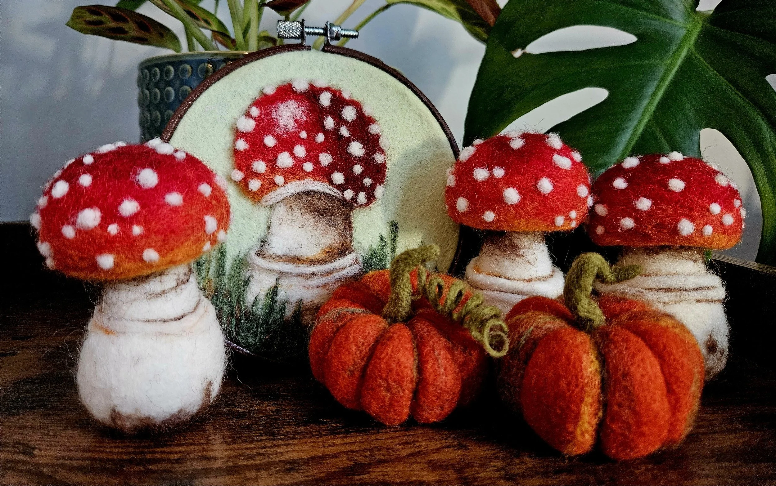 Felt mushroom and pumpkin decorations on a wooden surface, with a embroidered mushroom artwork and a leafy plant in the background.