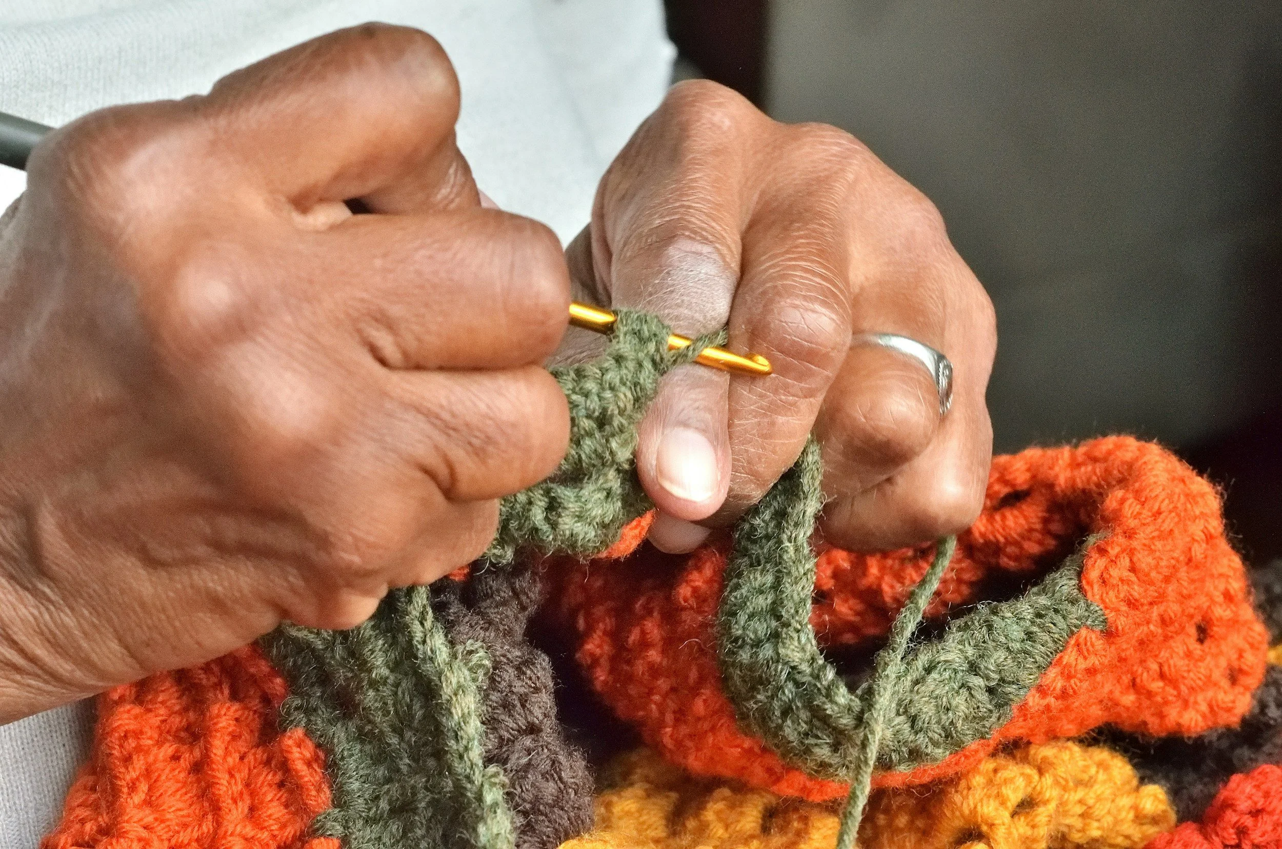 Close-up of hands knitting with green yarn on a gold knitting needle, with colorful knitted fabric in the background.