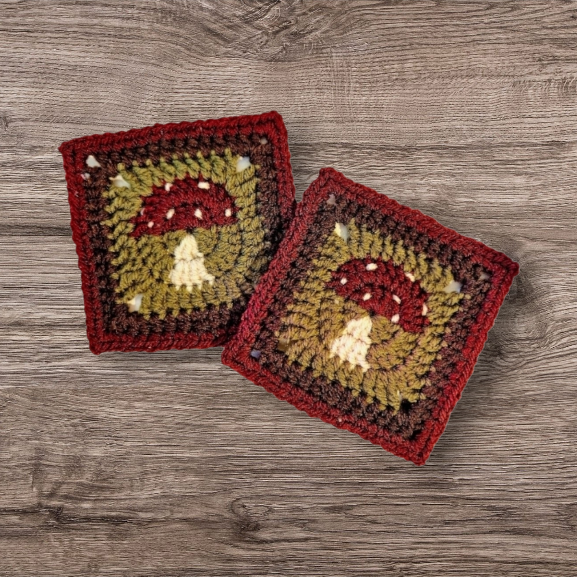 Two crocheted square patches with mushroom designs, featuring red caps with white spots and beige stems, bordered by red and dark brown yarn, on a wooden surface.