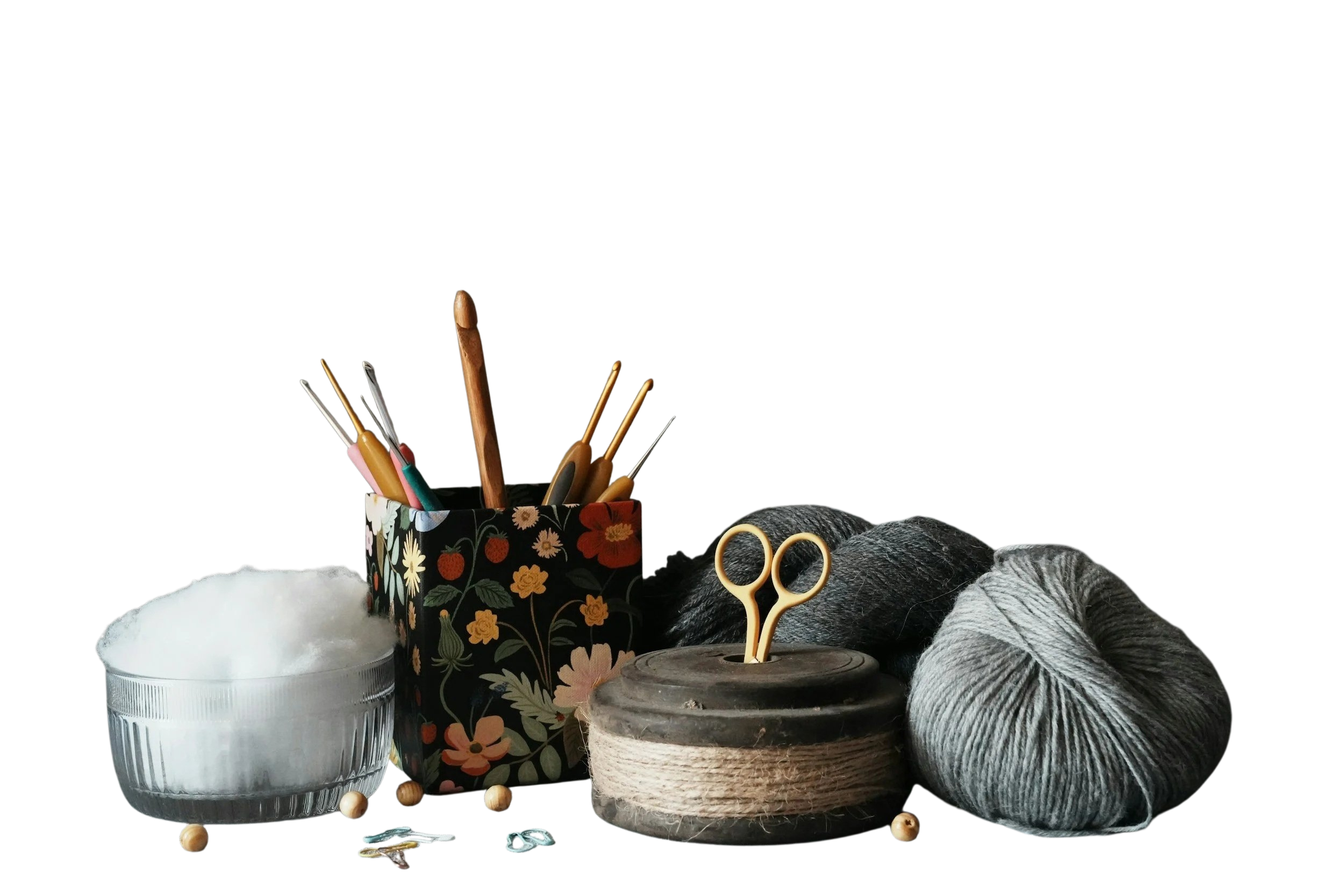 A collection of sewing and knitting supplies including yarn, scissors, a decorative container with pens and scissors, a bowl of cotton batting, a spool of thread, and some wooden sewing pins, set against a black background.