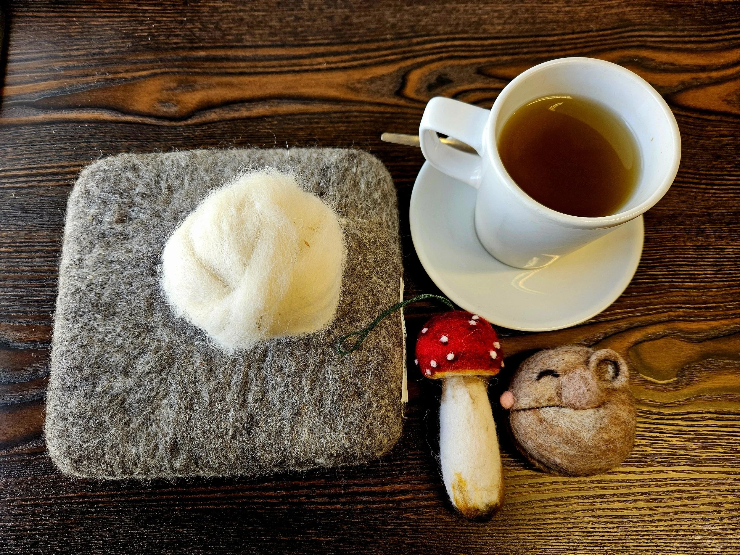 A felted wool mouse, a felted mushroom, and a felted mouse face with a cup of tea on a wooden table.