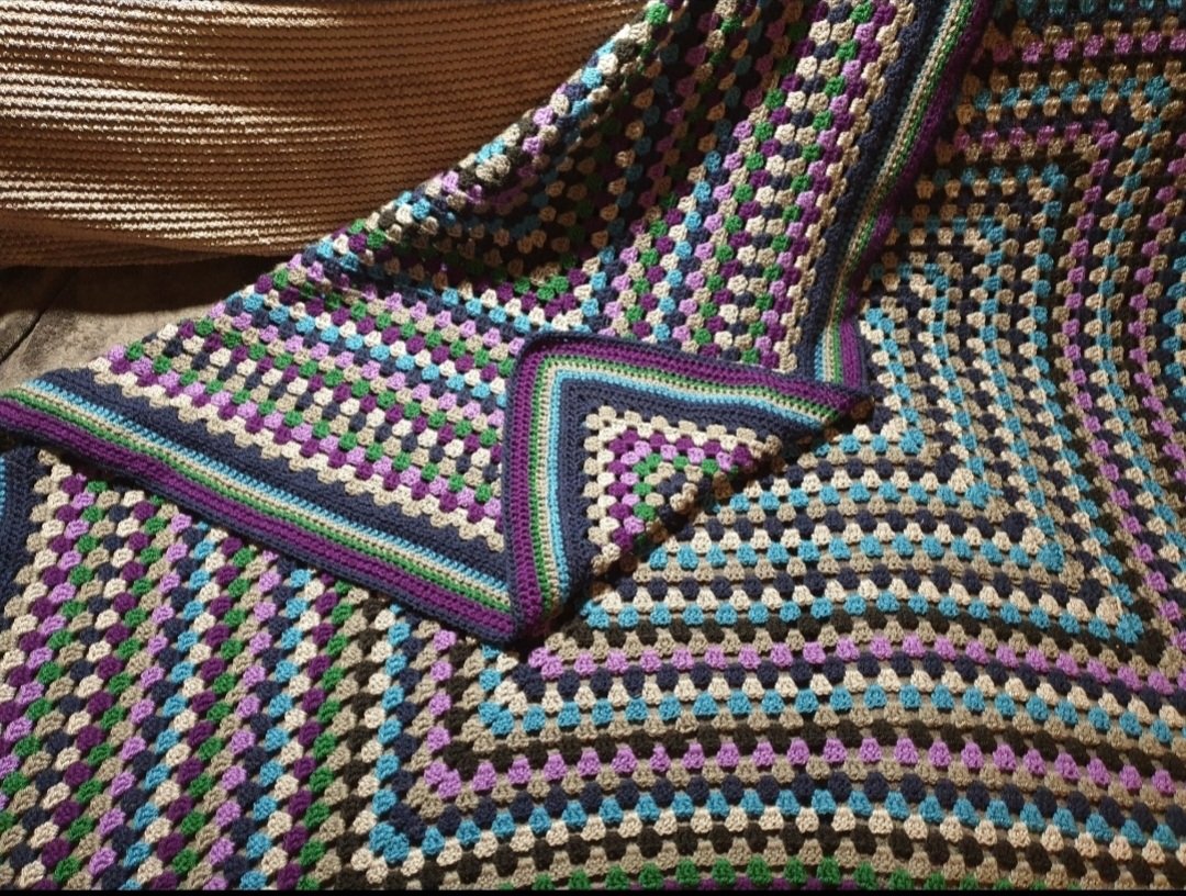 A colorful crocheted blanket with a geometric pattern in purple, green, blue, white, black, and beige, draped over a surface.