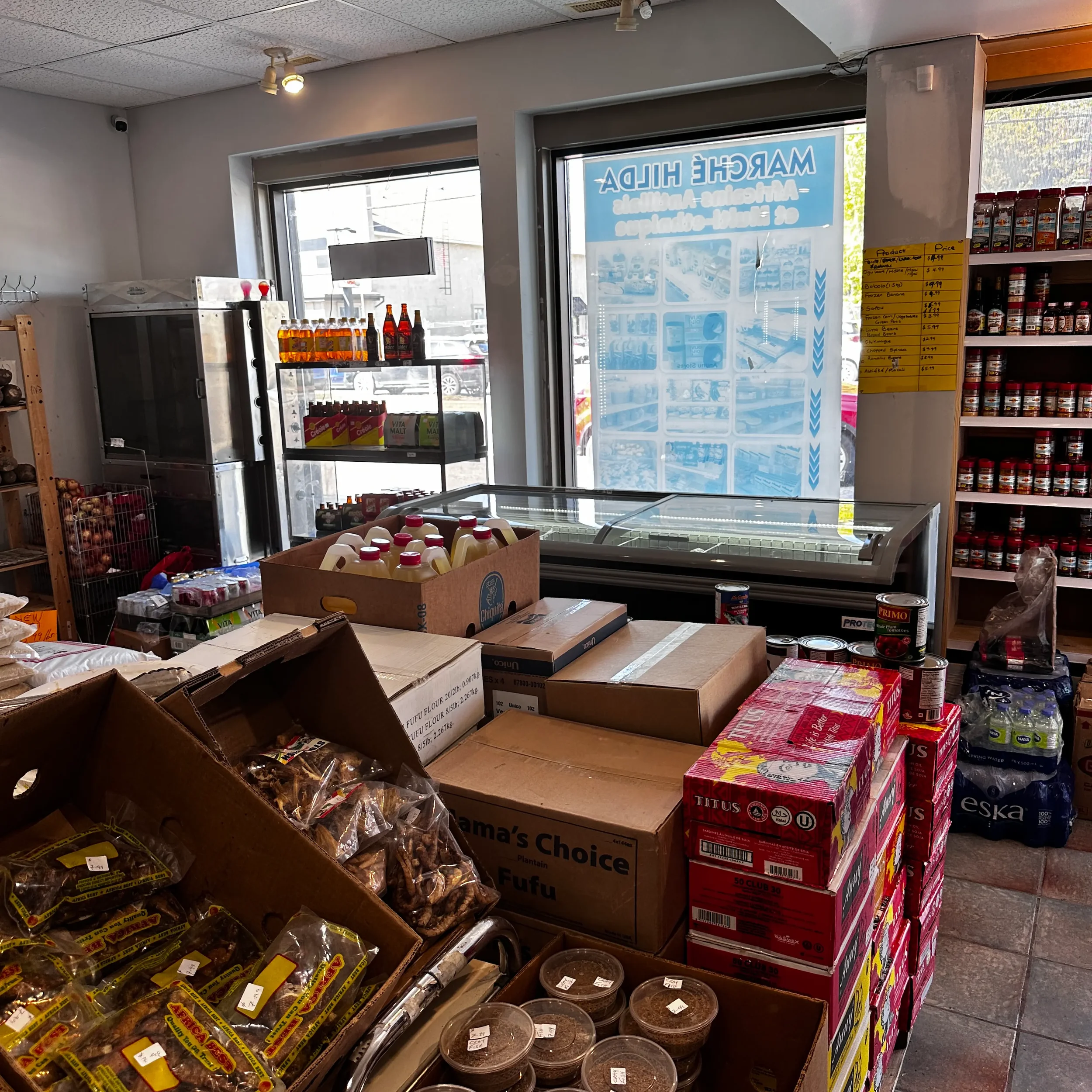 Inside a grocery store with various food and beverage items, including bottled drinks, boxed goods, and packaged snacks, near a refrigerated display case and shelves with jars, under store lighting.