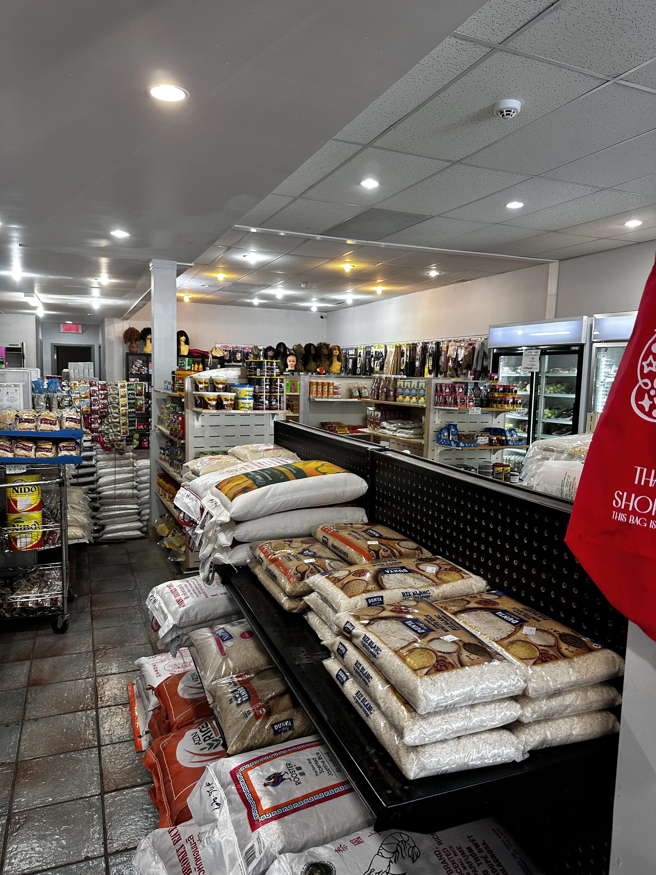 Interior of a grocery or convenience store with shelves stocked with rice, beans, and food products, and hats displayed on mannequin heads along the back wall.
