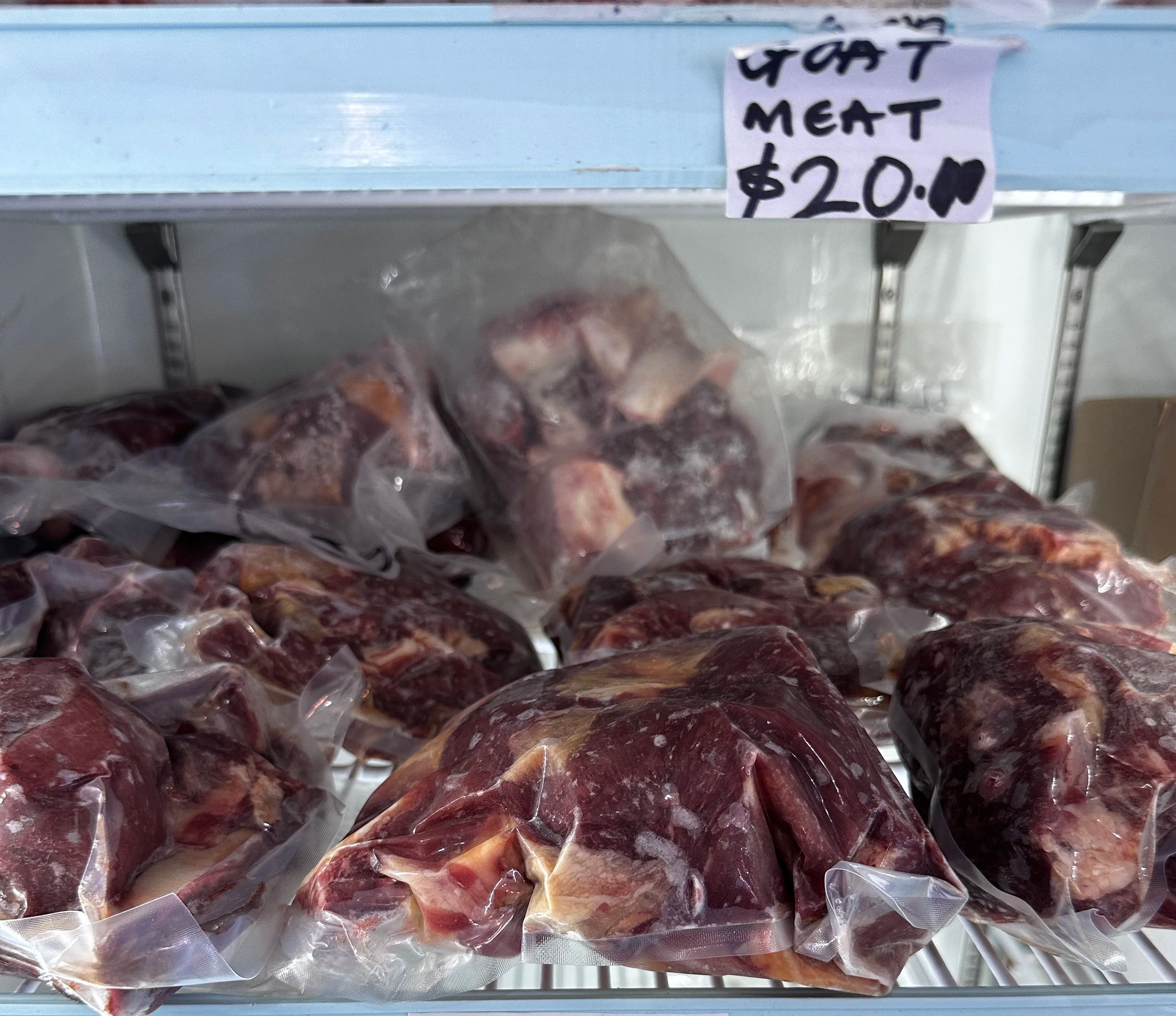 Frozen packages of goat meat on a grocery store shelf, with a handwritten sign indicating its price as $20 per pound.