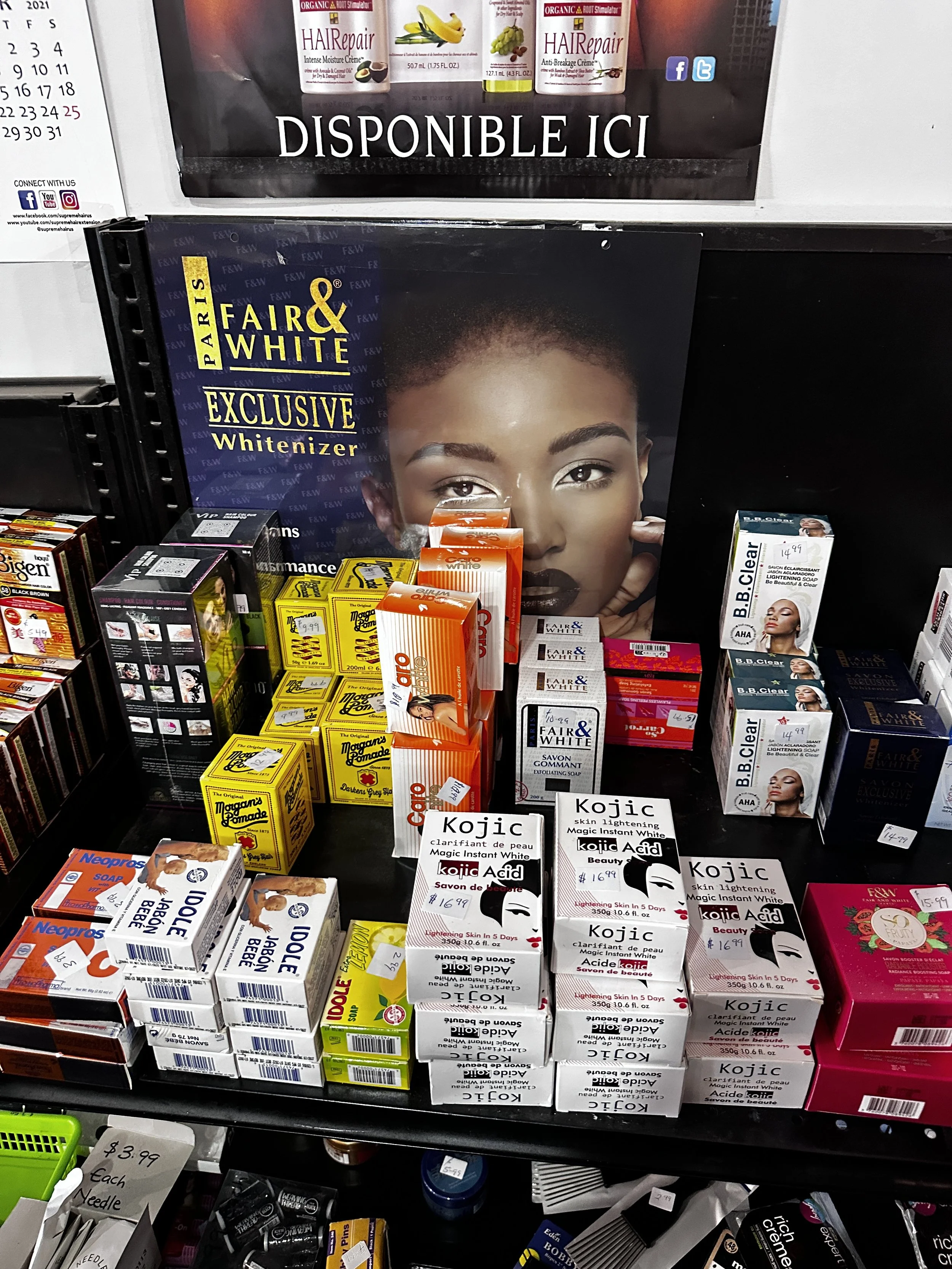 Shelf of skincare and beauty products, including Kojic acid skin brightening cream, B.B. Clear skincare, and products by Fair & White, with promotional poster in the background showing a woman's face.