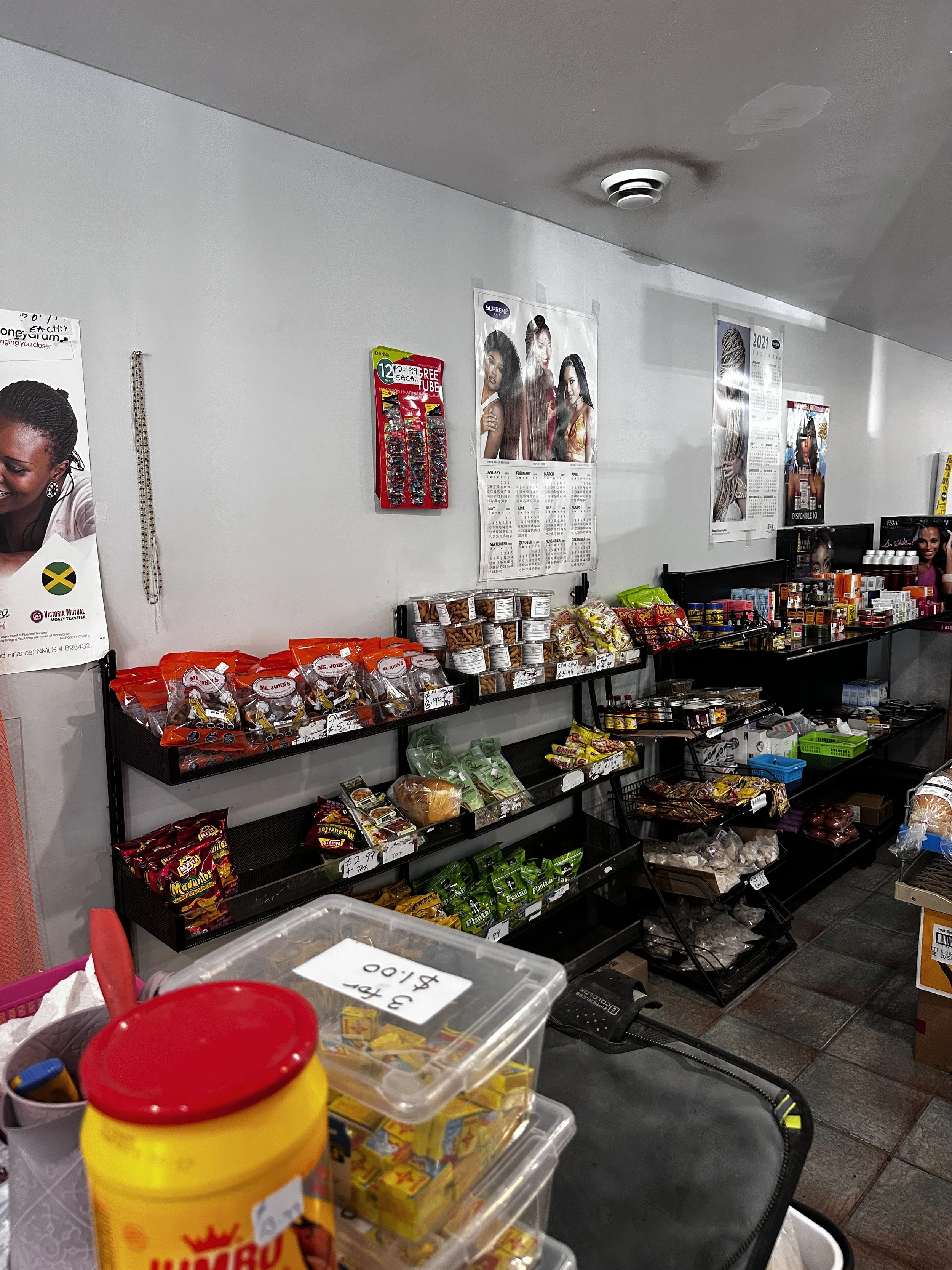 Interior of a small store with shelves stocked with snacks, sauces, and other food items. Posters of women with hairstyles cover the wall.
