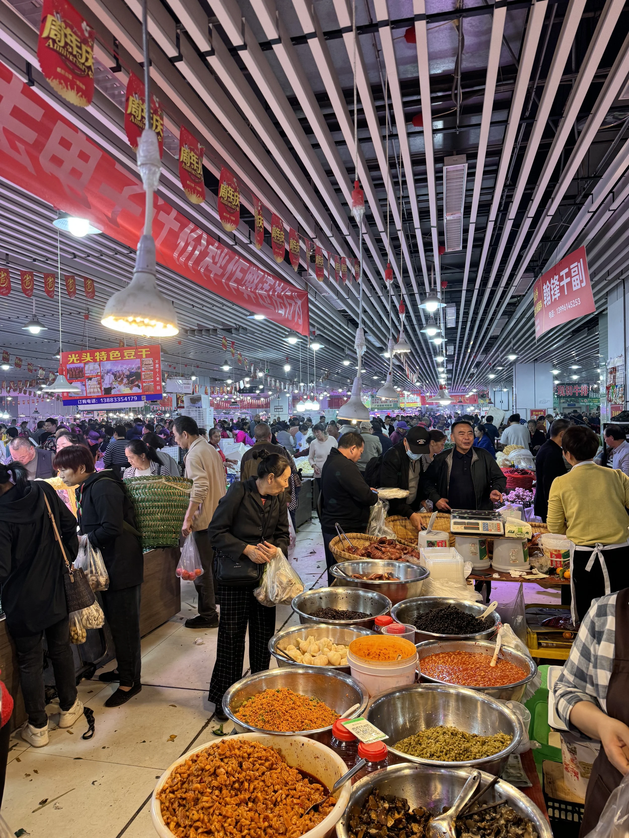 A bustling indoor market with people shopping and vendors selling various food items, including spices, dried goods, and prepared dishes.
