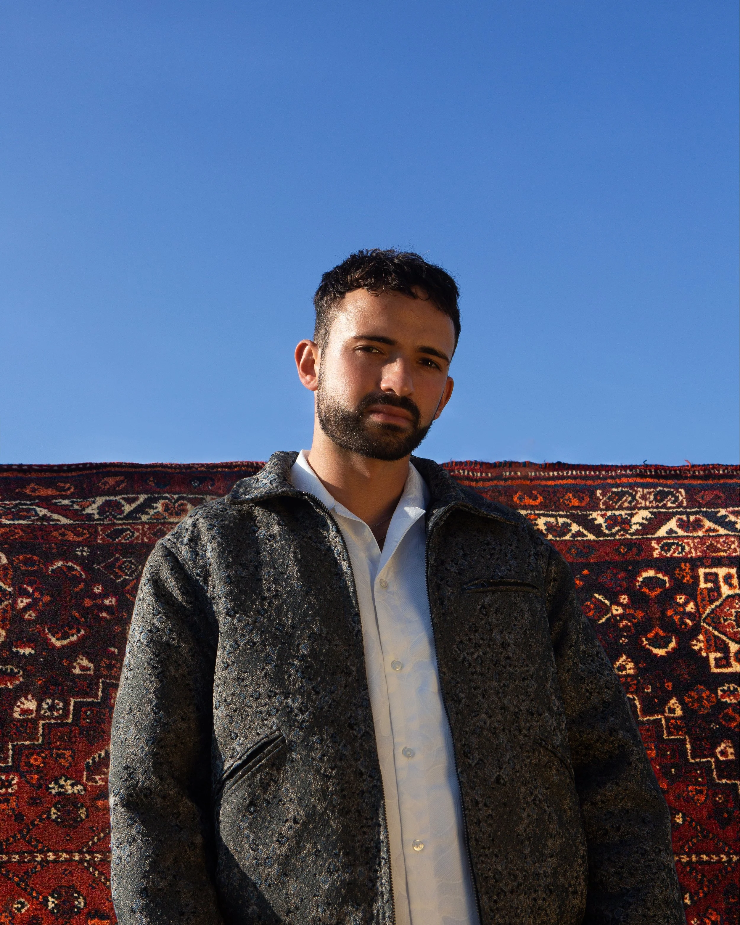 A man with dark hair and beard wearing a white shirt and patterned jacket standing outdoors with a colorful woven rug behind him and a clear blue sky.