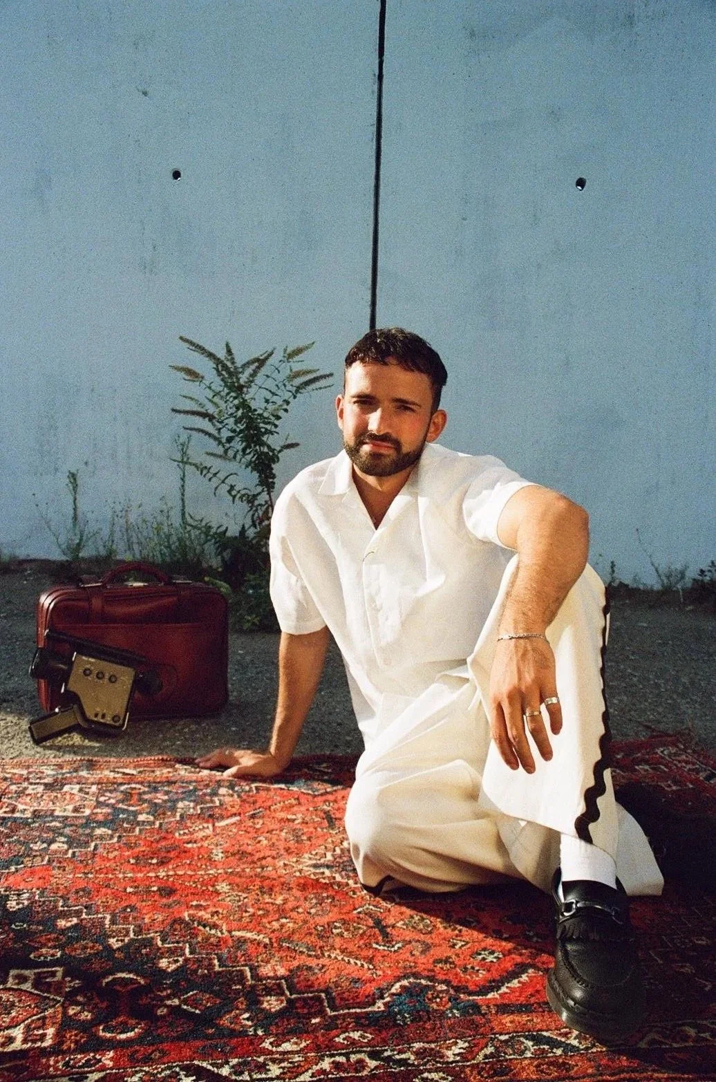 A man with a beard and short hair, dressed in white, sitting on a patterned rug against a blue wall with small plants. There is a red suitcase and a vintage record player nearby.
