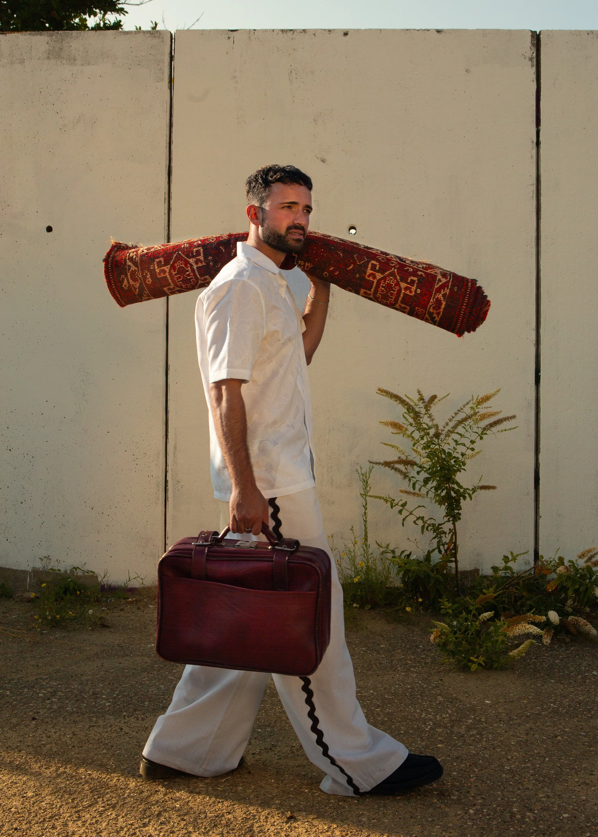 A man with a beard in white clothes walks outside carrying a maroon briefcase in one hand and has a rolled rug on his shoulder.