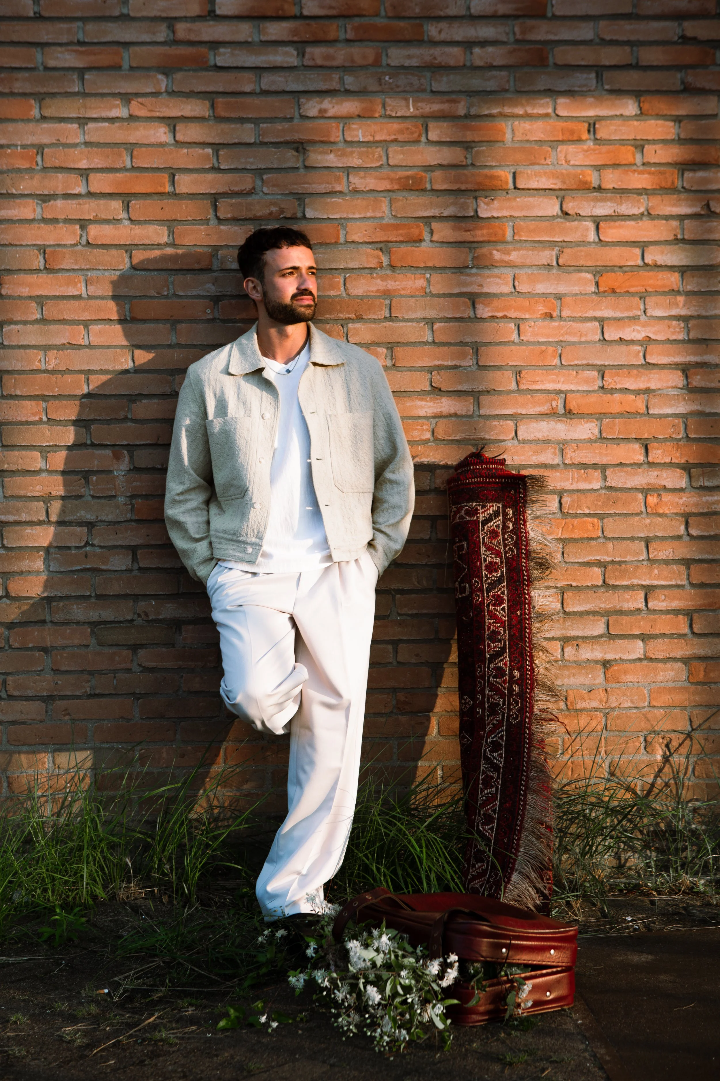 A man with a beard stands leaning against a brick wall, dressed in a light-colored jacket and white pants. There is a rolled-up rug, a brown suitcase, and a bouquet of white flowers on the ground nearby, with tall grass and dirt in the foreground.