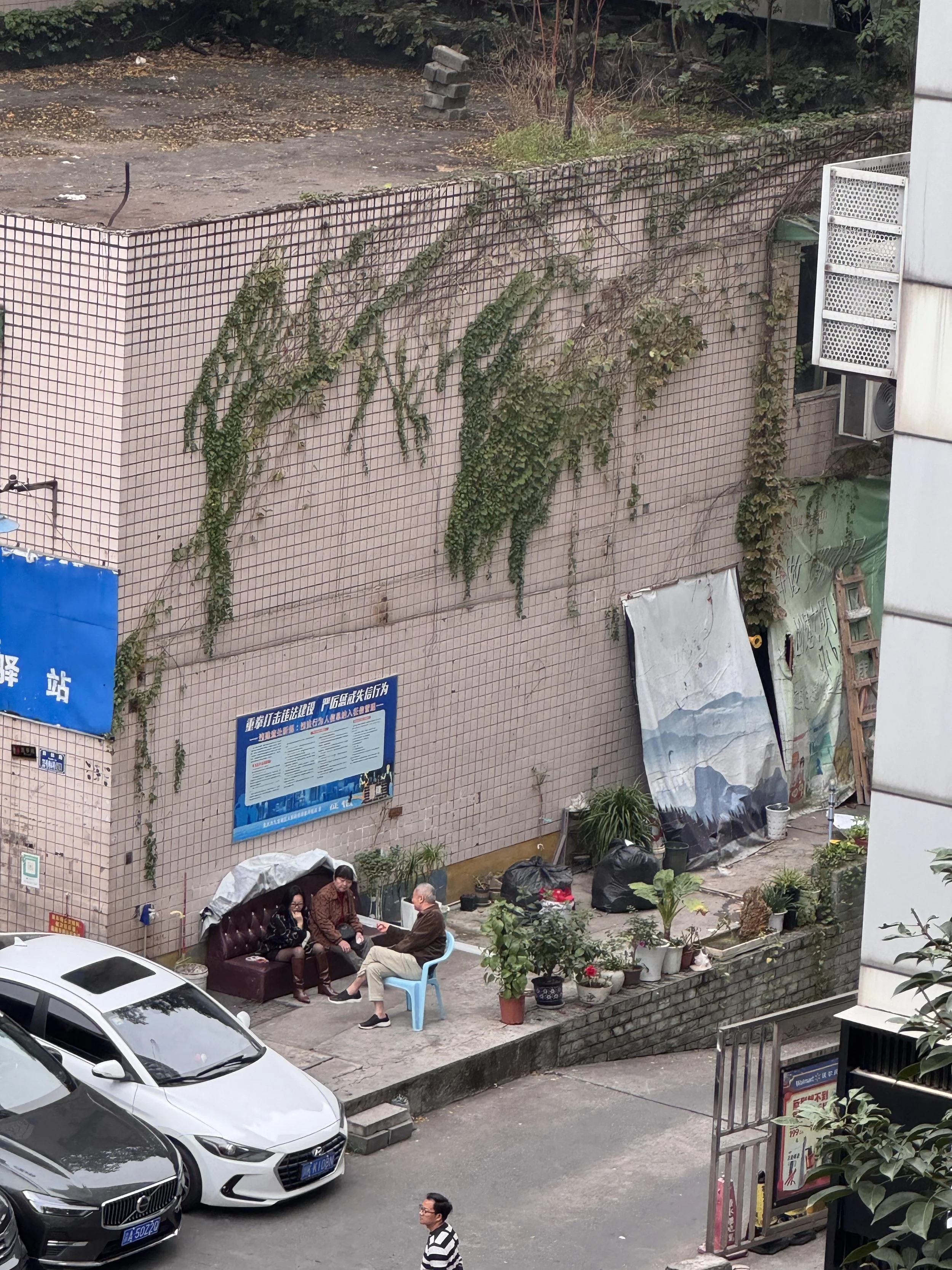 Three people sitting on a small sidewalk corner, having a conversation. There are cars parked nearby, potted plants, a mural, and various items around, with a pink tiled wall in the background overgrown with vines.