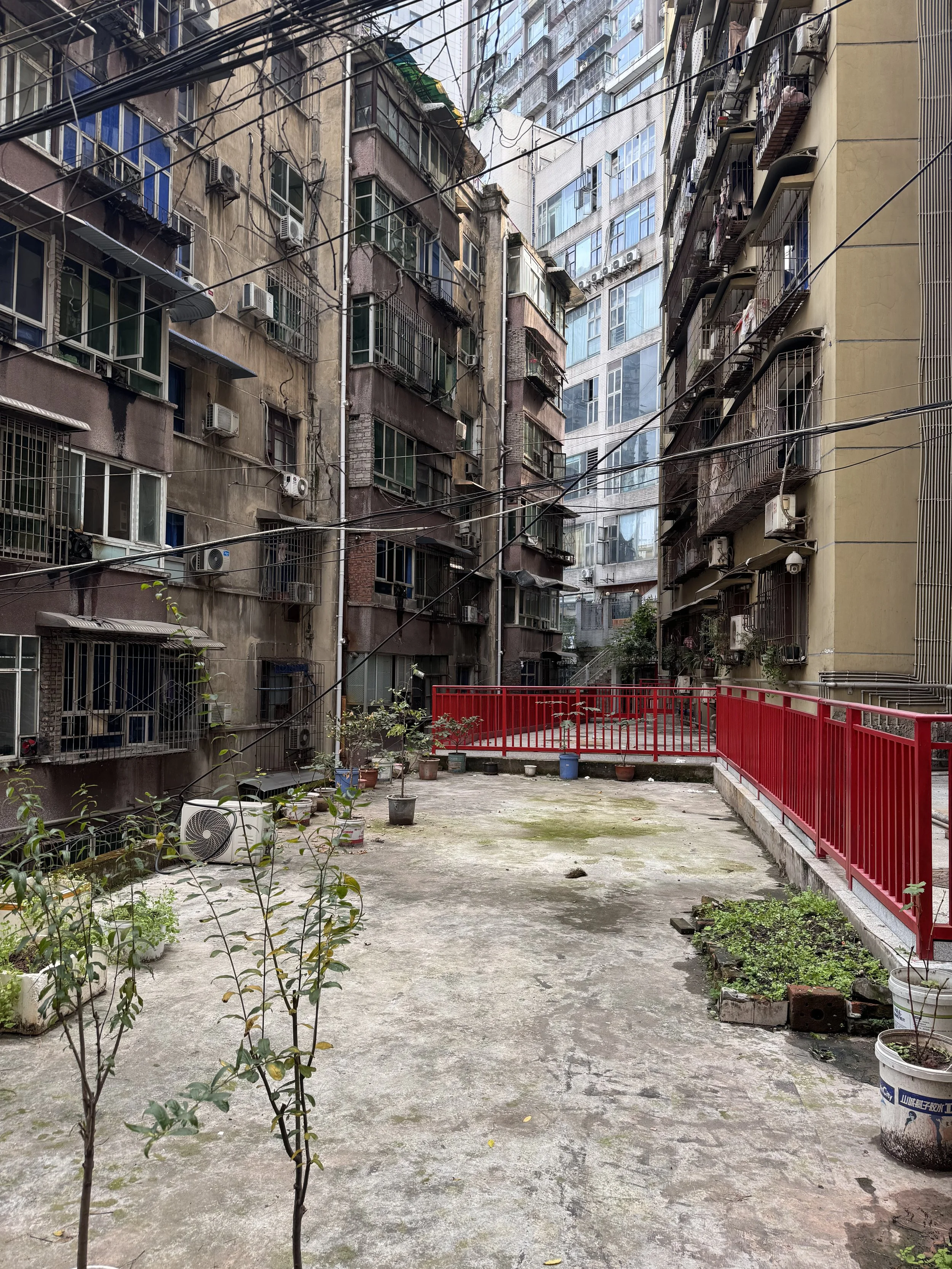 An urban courtyard surrounded by tall, weathered apartment buildings with numerous windows, some with air conditioning units. The courtyard has potted plants, a concrete surface with patches of moss, and a bright red railing along one edge, creating a stark contrast against the worn buildings.