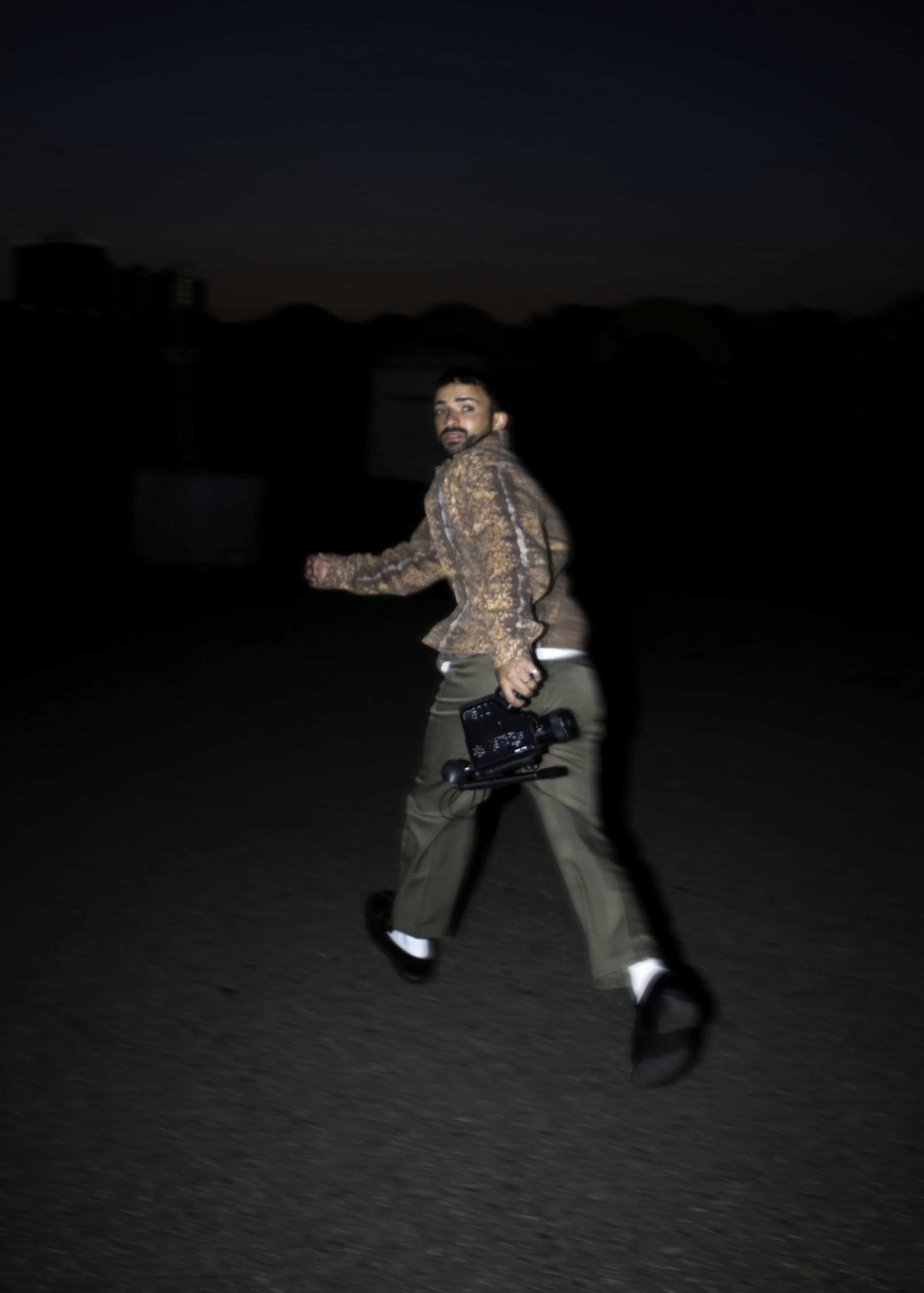 A man walking outdoors at night, holding a camera in his right hand, wearing a brown patterned jacket and khaki pants.