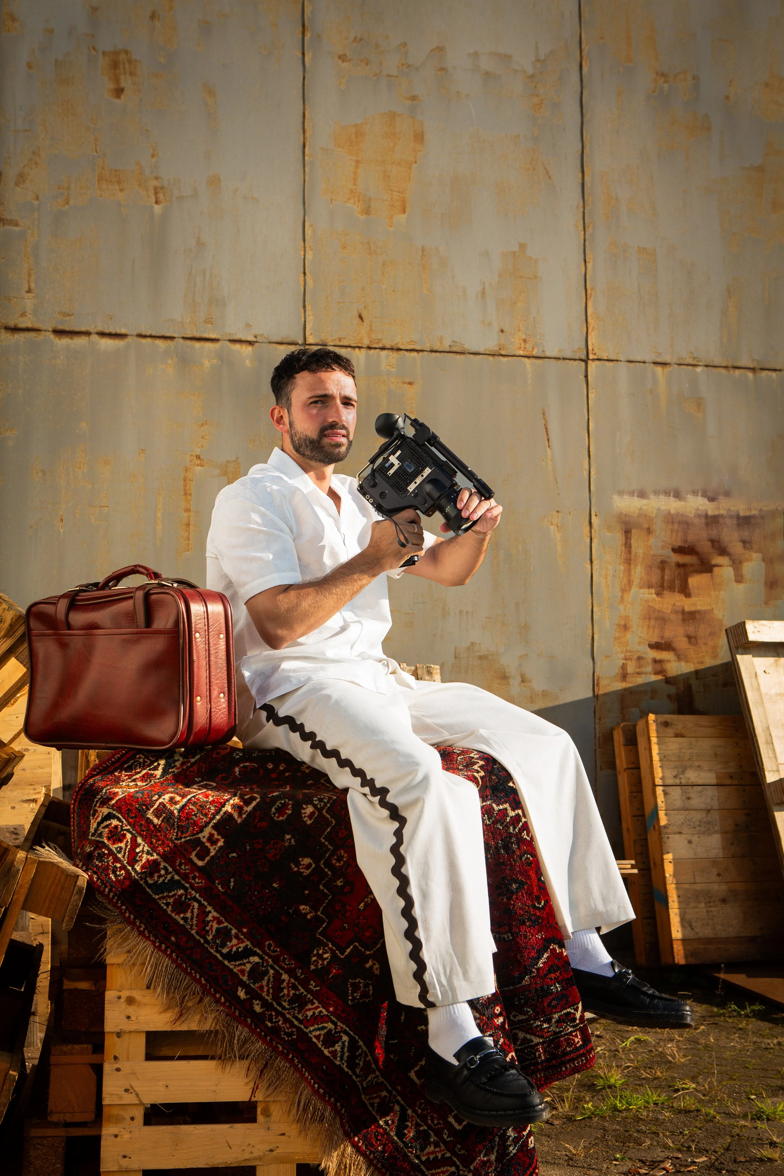 A man dressed in white sitting on a wooden crate, holding a video camera, with a patterned rug underneath, a brown leather bag next to him, and a rustic metal wall in the background.