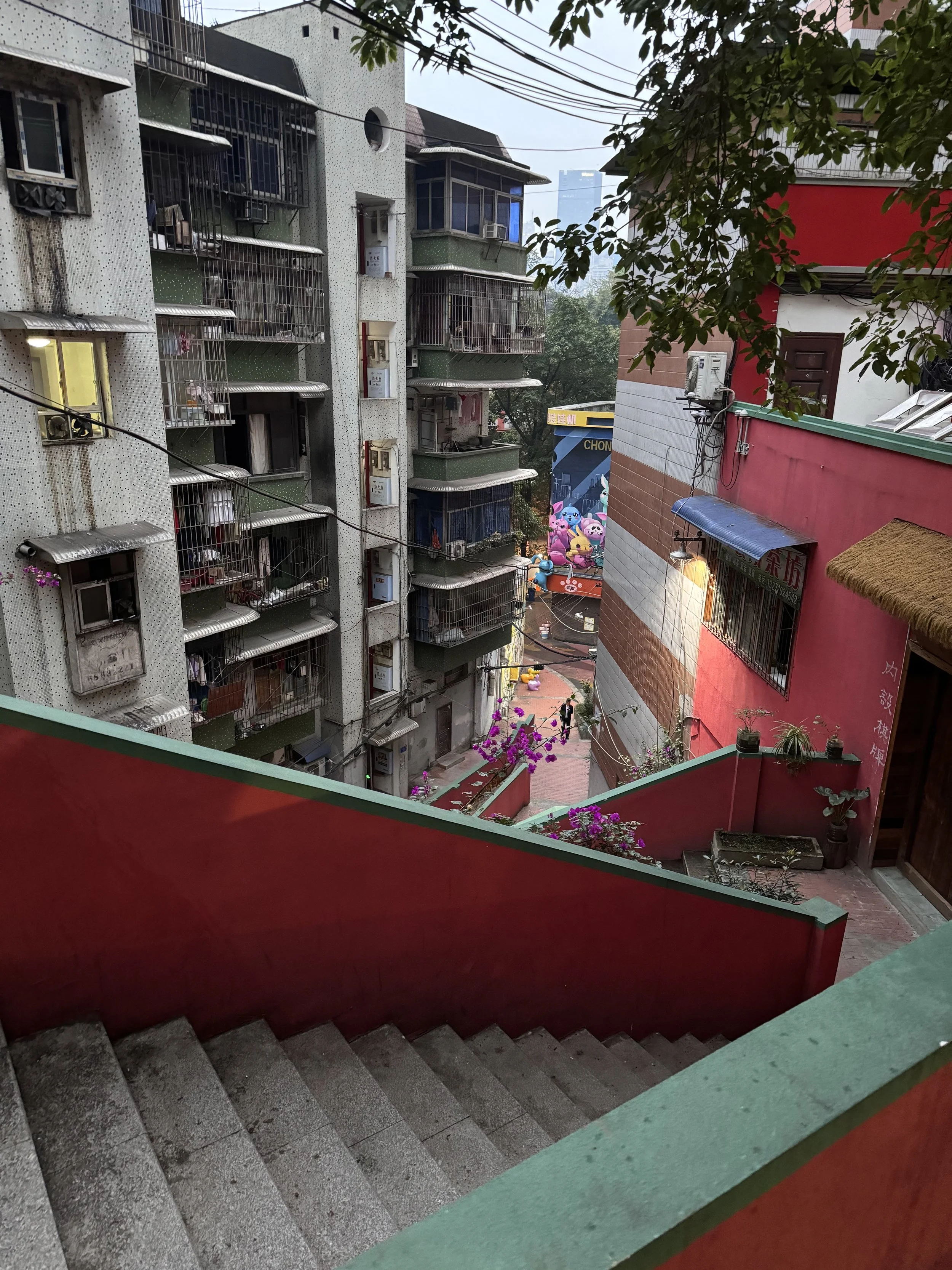 View from a staircase looking down a city alley with multi-story residential buildings, colorful walls, potted plants, and a decorated outdoor area with pink and purple balloons.