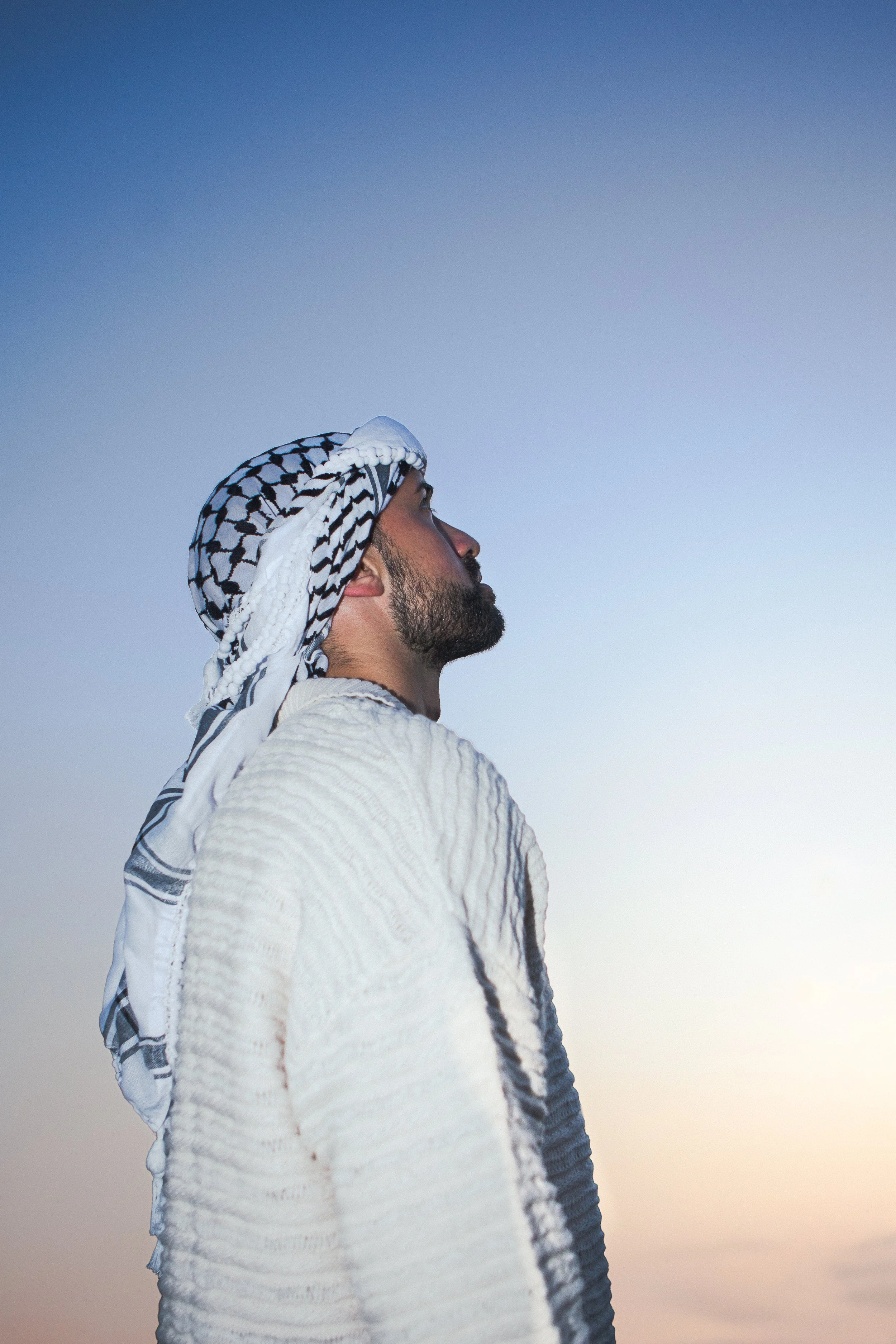 A man with a beard wearing a traditional Arab head covering and a white textured robe, standing sideways and looking up against a clear blue sky at sunset.