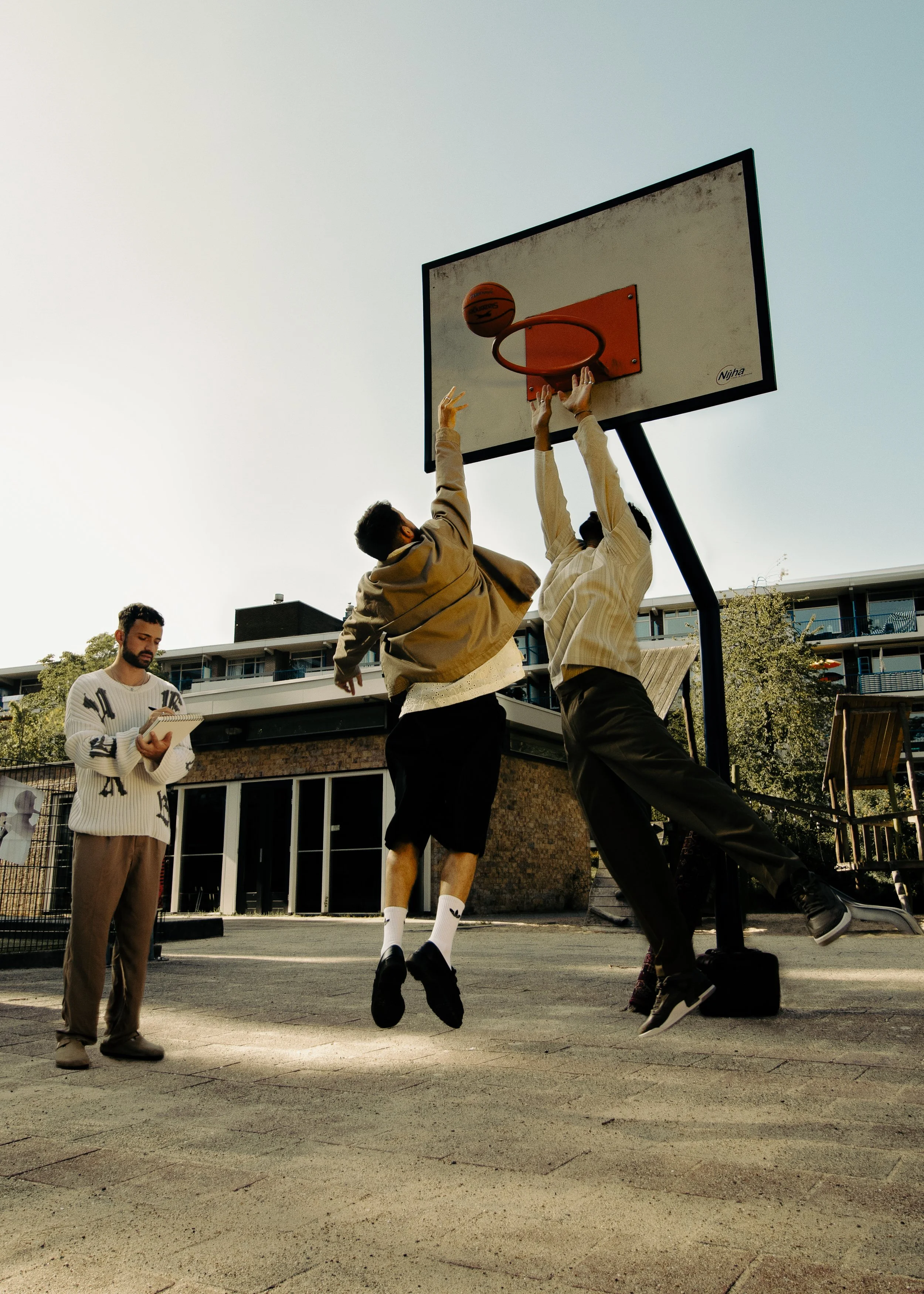 Two young men playing basketball on an outdoor court, one attempting to make a shot while the other tries to block, with a third man observing and taking notes nearby.